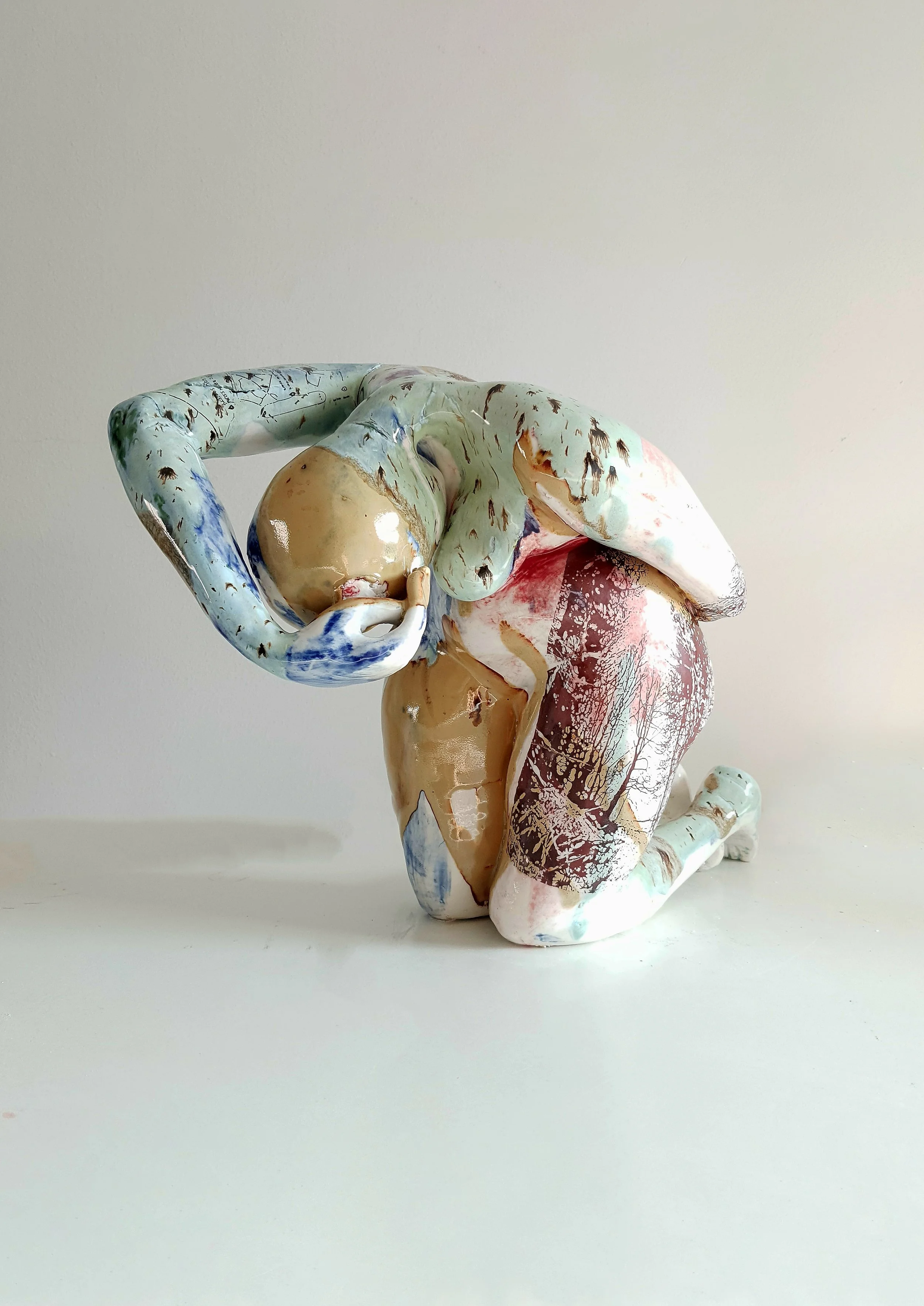 A colorful ceramic sculpture of a person kneeling with their head resting in their hands, portrayed in a crouched position.