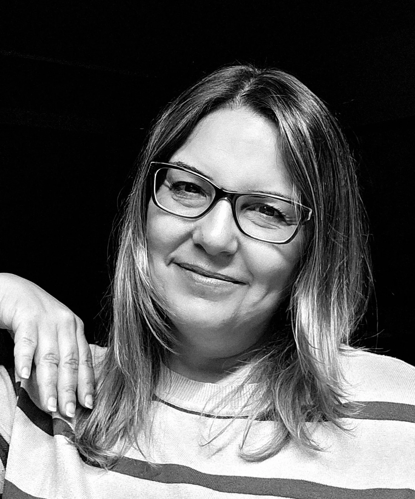 Black and white portrait of a smiling woman with glasses and shoulder-length hair, wearing a striped shirt.