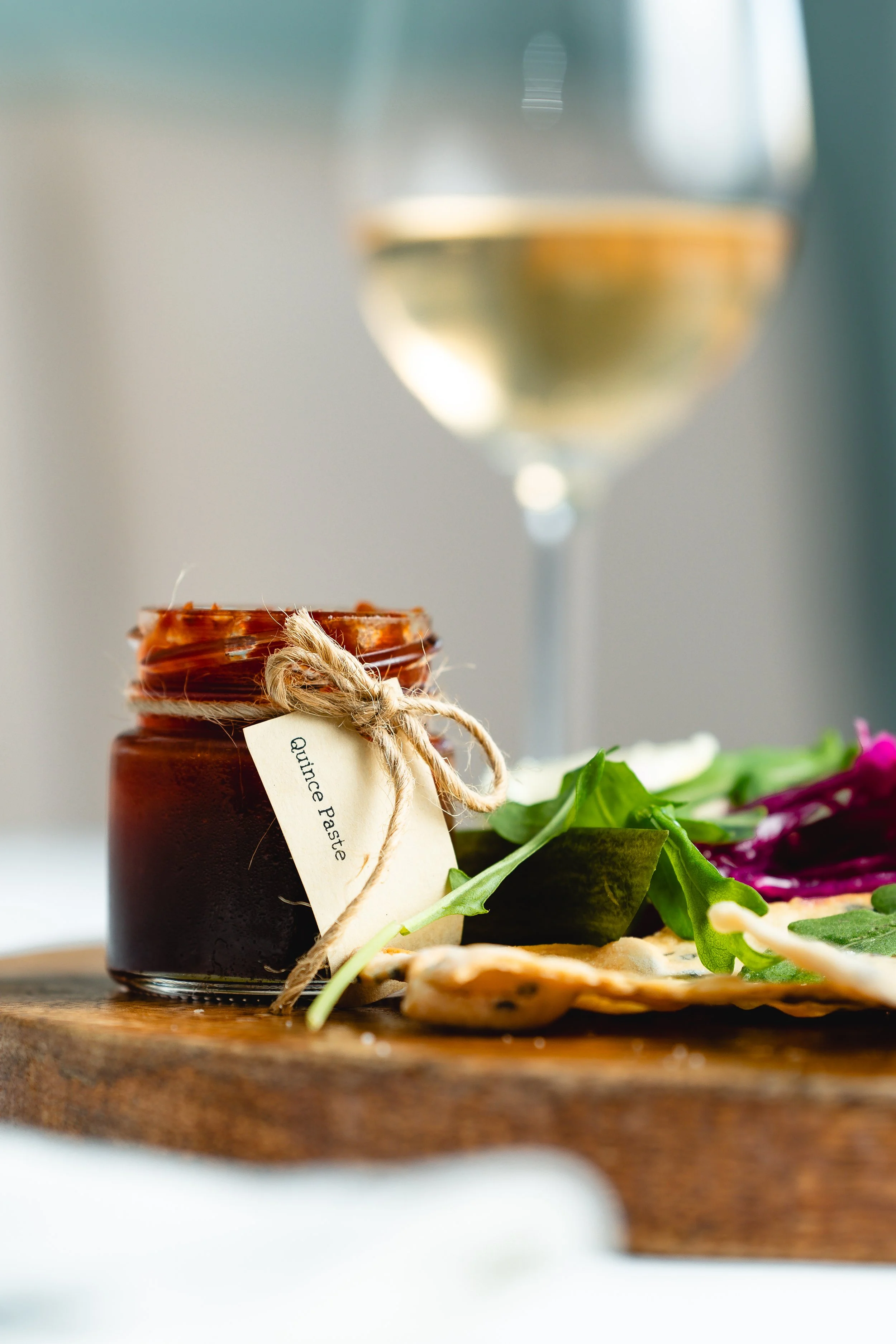A small jar labeled Quince Paste tied with a string, placed on a wooden board with leafy greens, accompanied by a glass of white wine in the background.