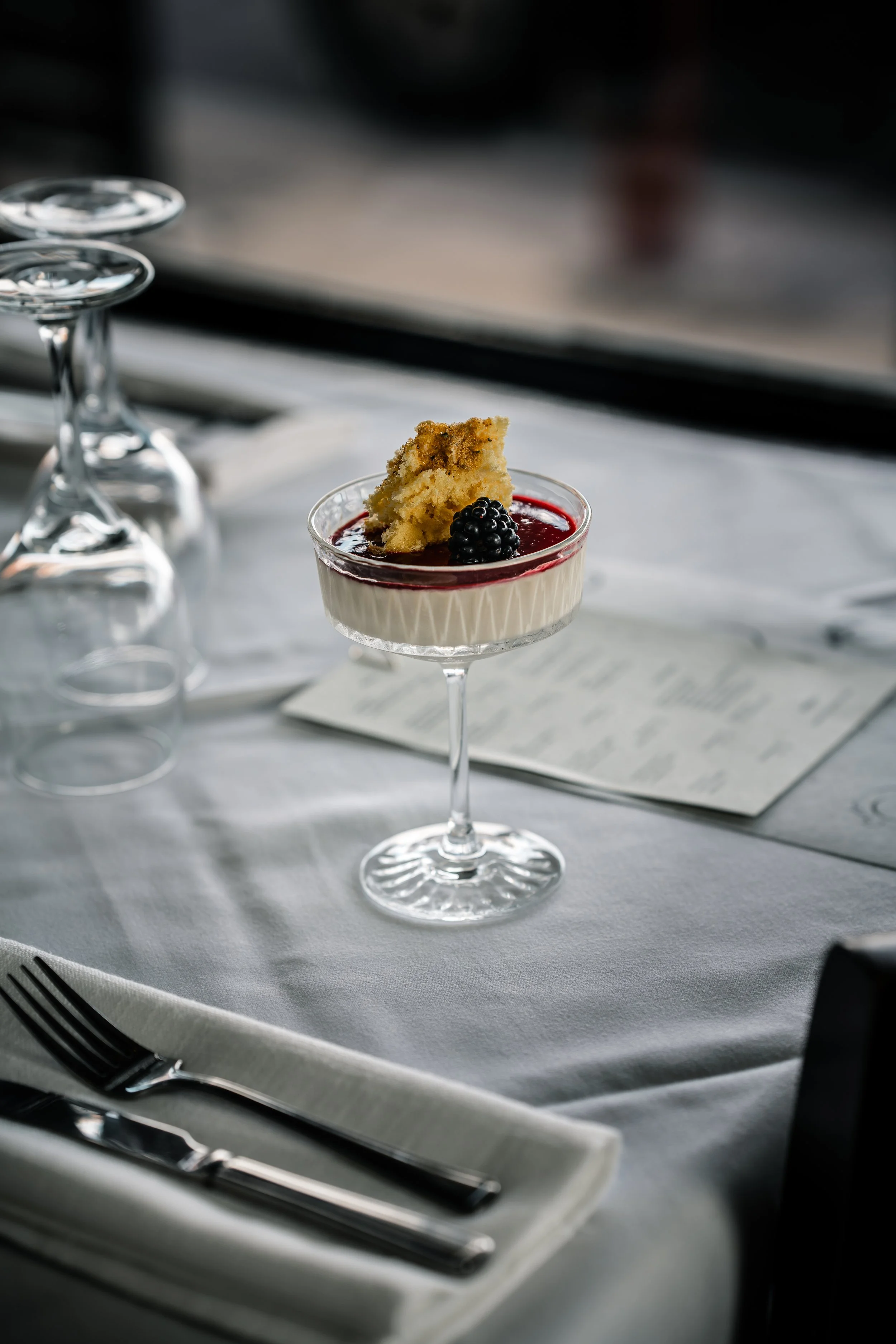 A dessert in a glass on a white tablecloth with a fork and knife nearby, featuring a slice of cake, blackberries, and a red sauce topping.