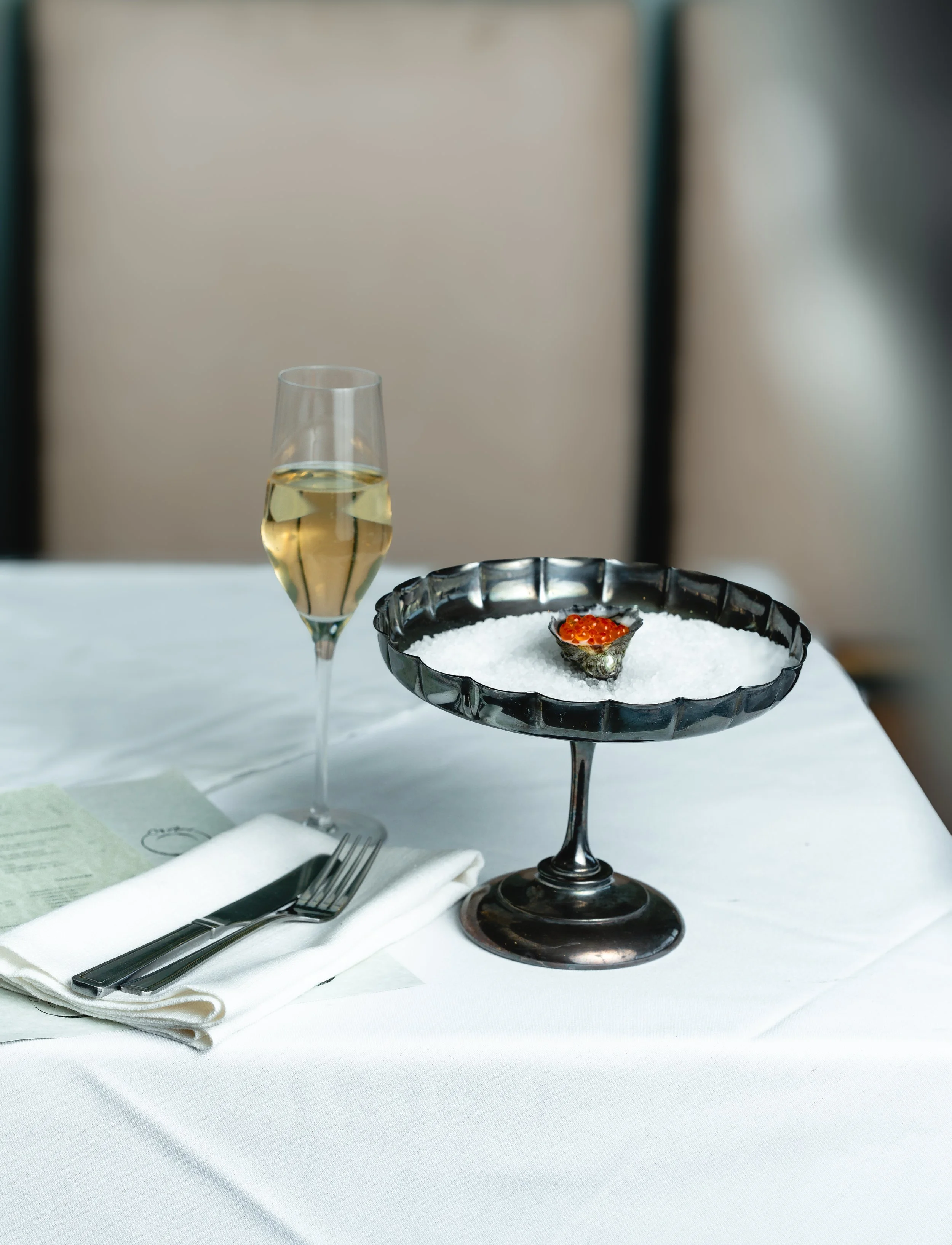 A glass of white wine, a fork and knife on a white napkin, and a dish of salmon roe on a bed of ice on a white tablecloth.