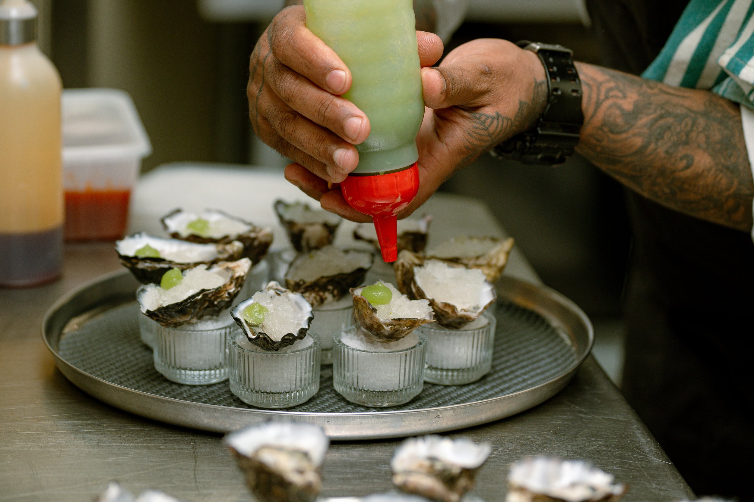 A chef or worker with tattooed arms is squeezing a bottle of sauce over oyster shells filled with ice and topped with a white creamy substance and green garnish, on a tray.