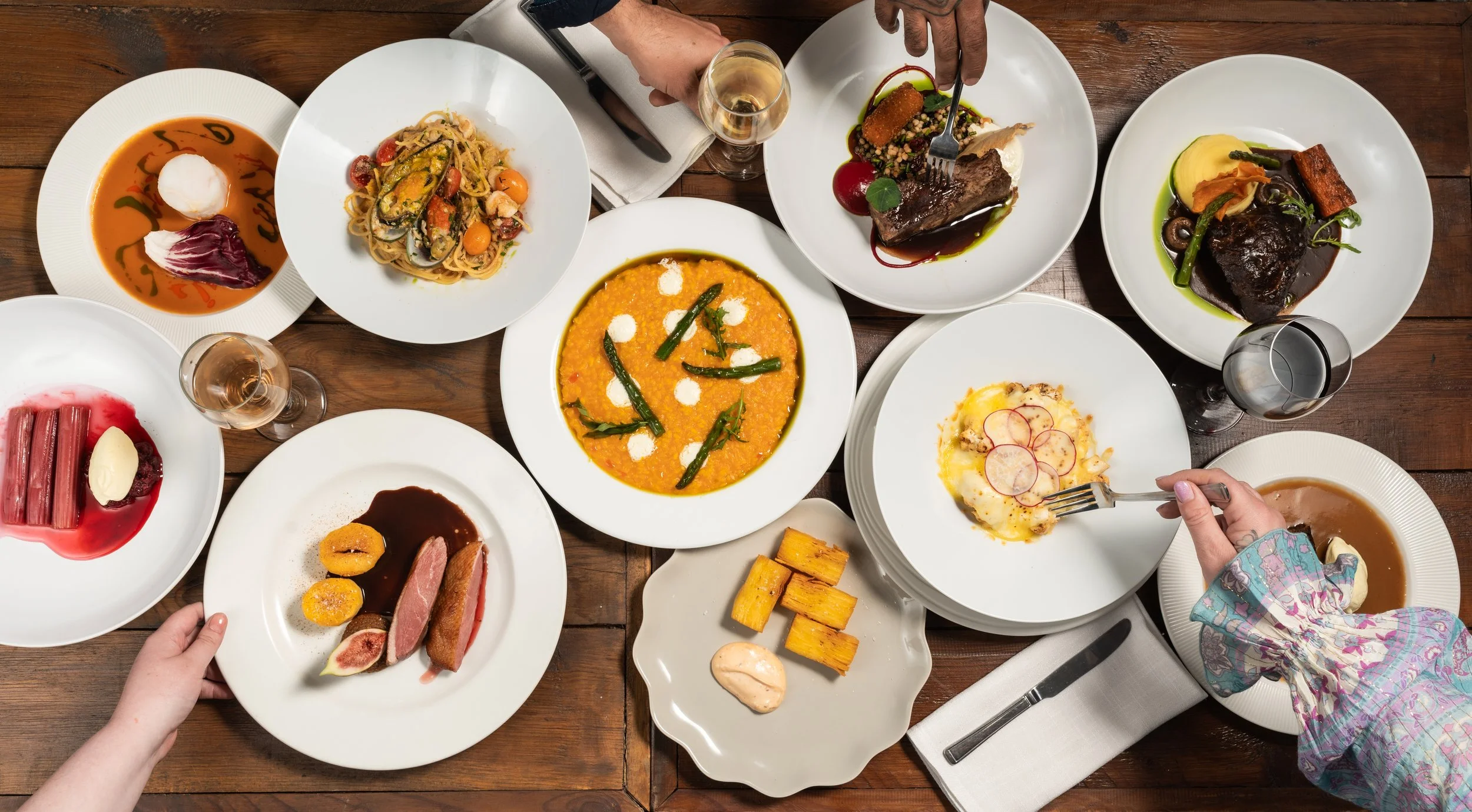 A table filled with various plates of gourmet food, including soups, pasta, meat dishes, desserts, and glasses of white wine, set on a wooden table with some hands reaching for the dishes.