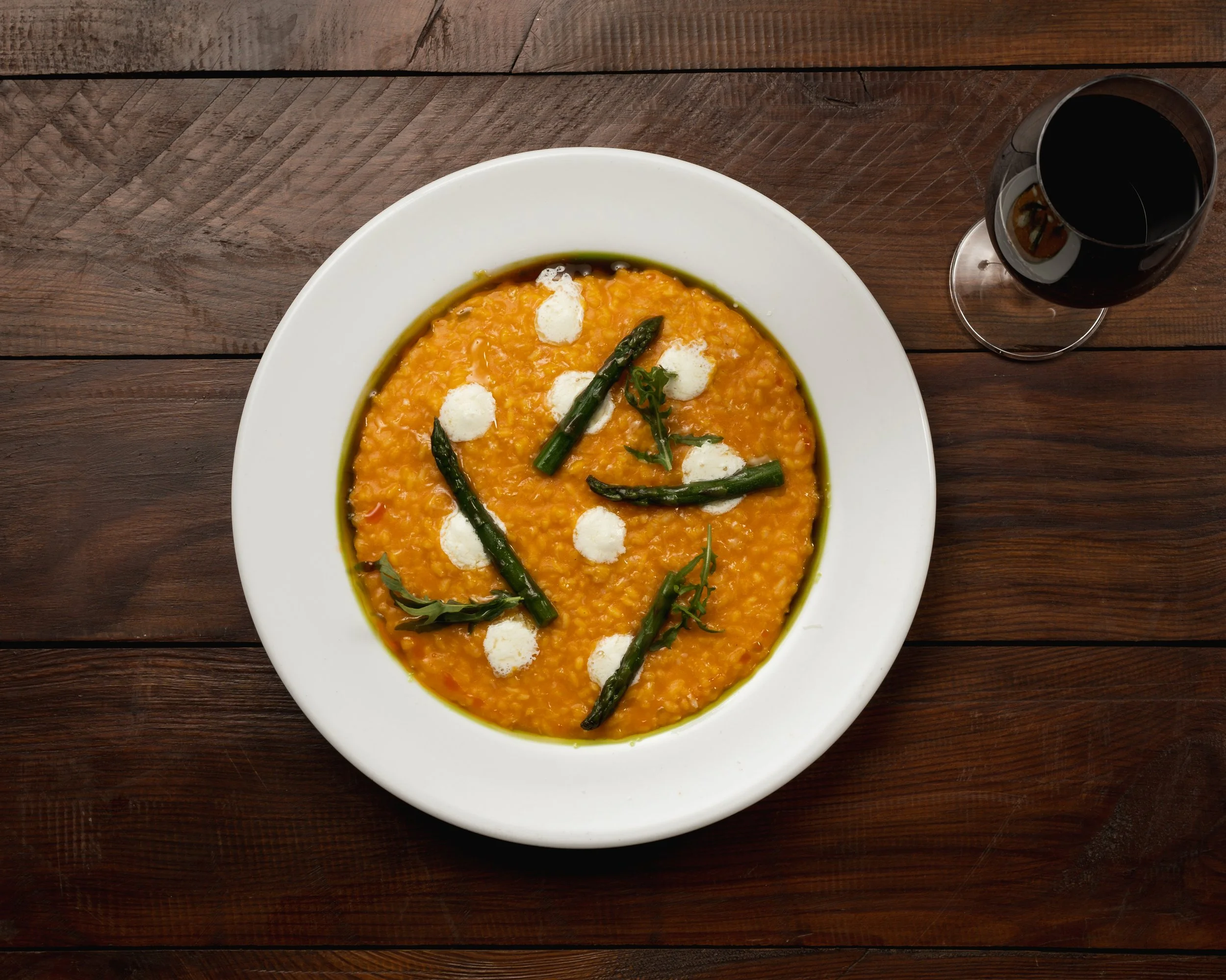 A bowl of orange lentil soup garnished with green beans and dollops of white sauce or cream, with a glass of red wine on a wooden table.