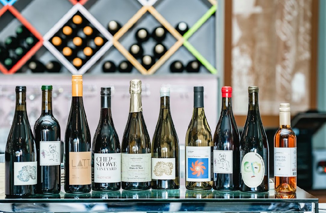A row of nine wine bottles with different labels, some with foil capsules, behind a glass bar counter in a wine store or bar.