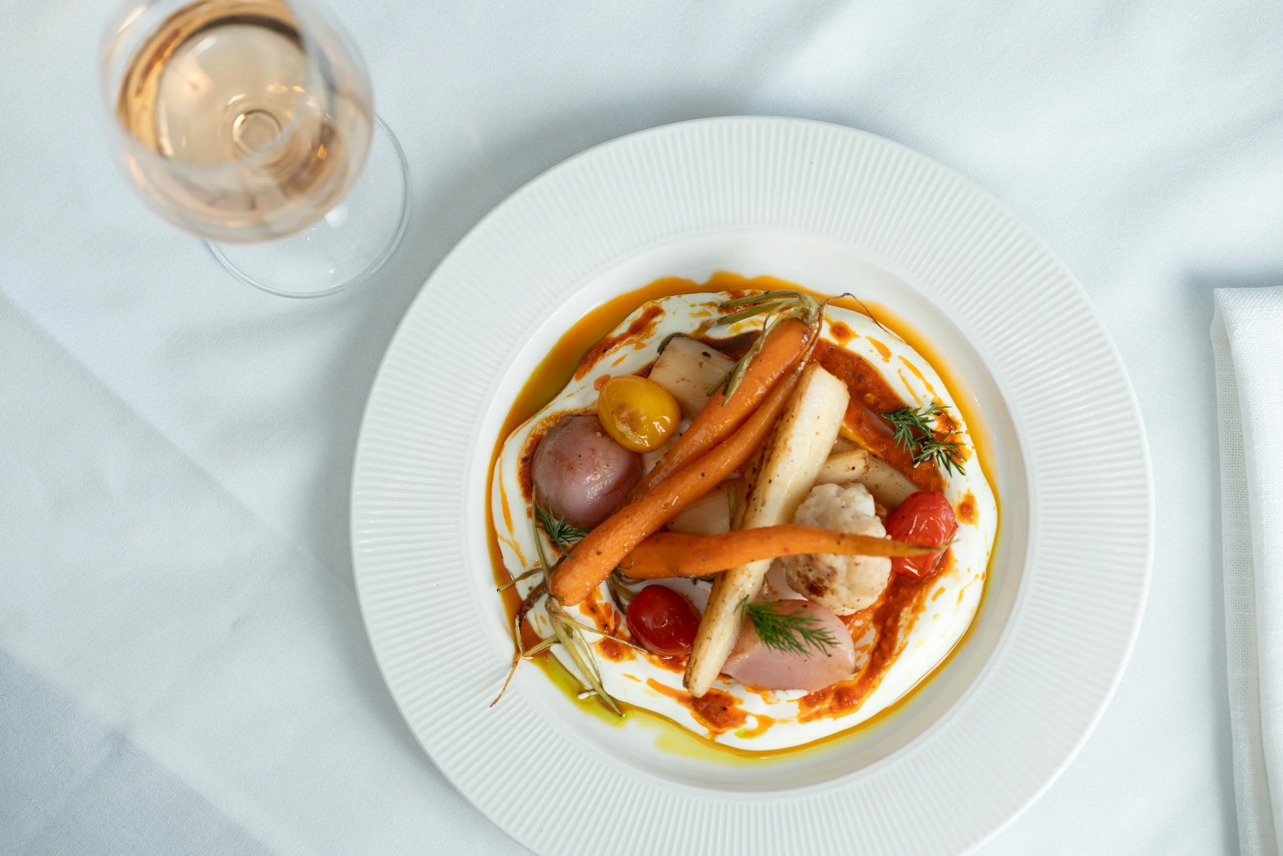A plate of assorted roasted vegetables and a glass of white wine on a white tablecloth.