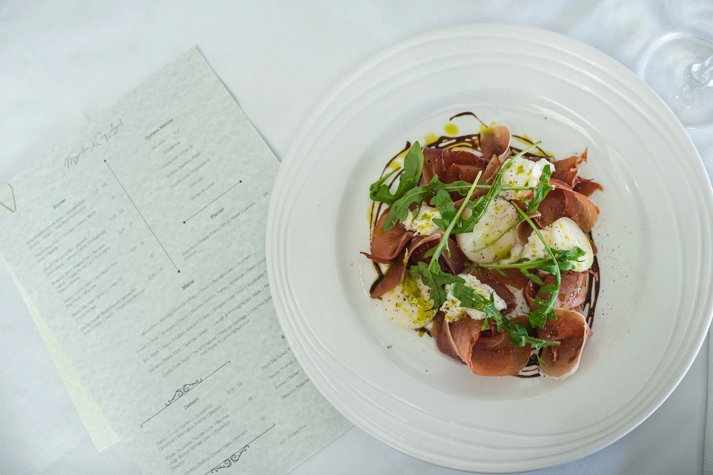 A dish of prosciutto, mozzarella, arugula, and balsamic glaze on a white plate, with a menu nearby and a glass of water in the background.