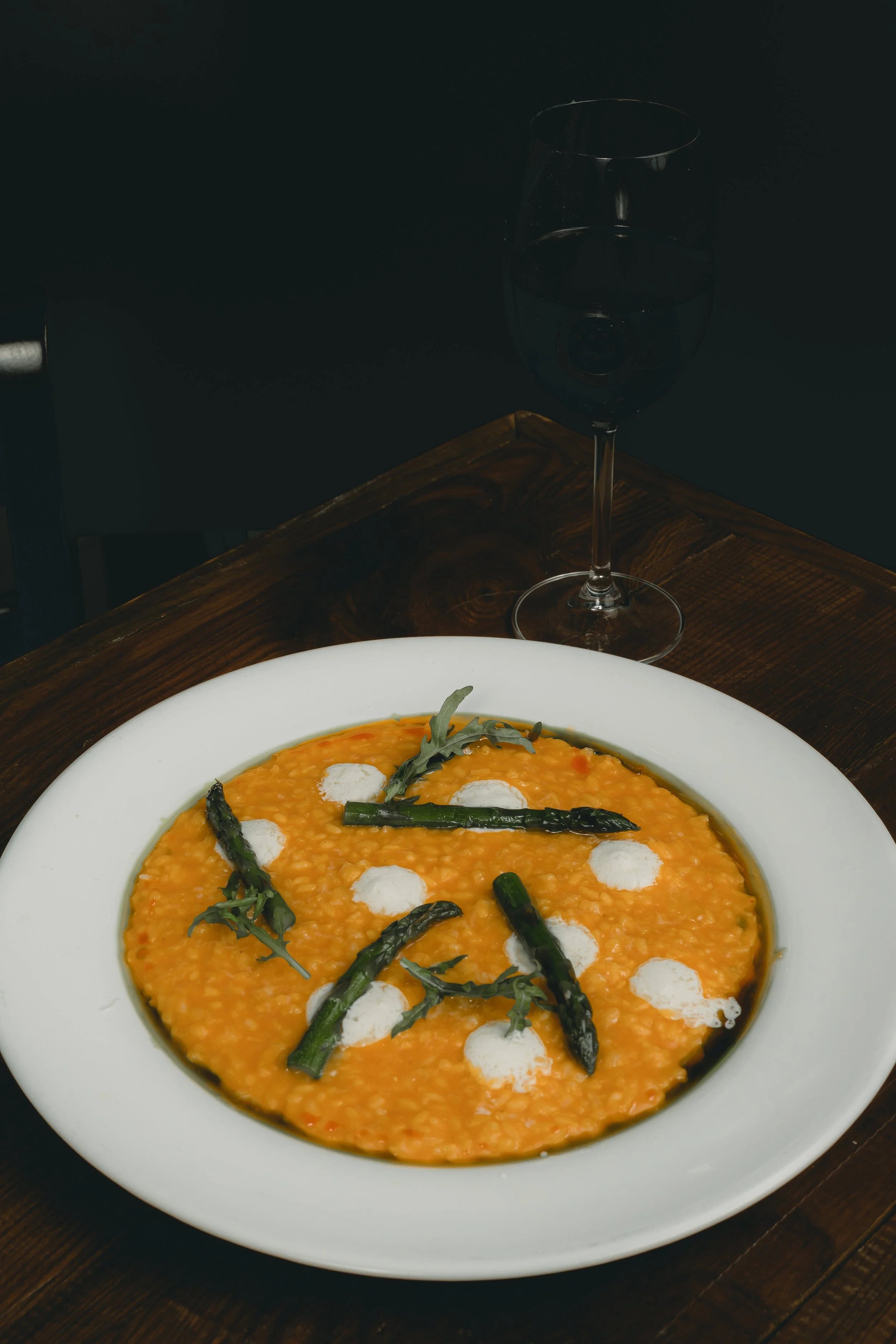 A white plate with orange soup garnished with green asparagus and small dollops of white cream, placed on a dark wooden table, with a glass of water in the background.