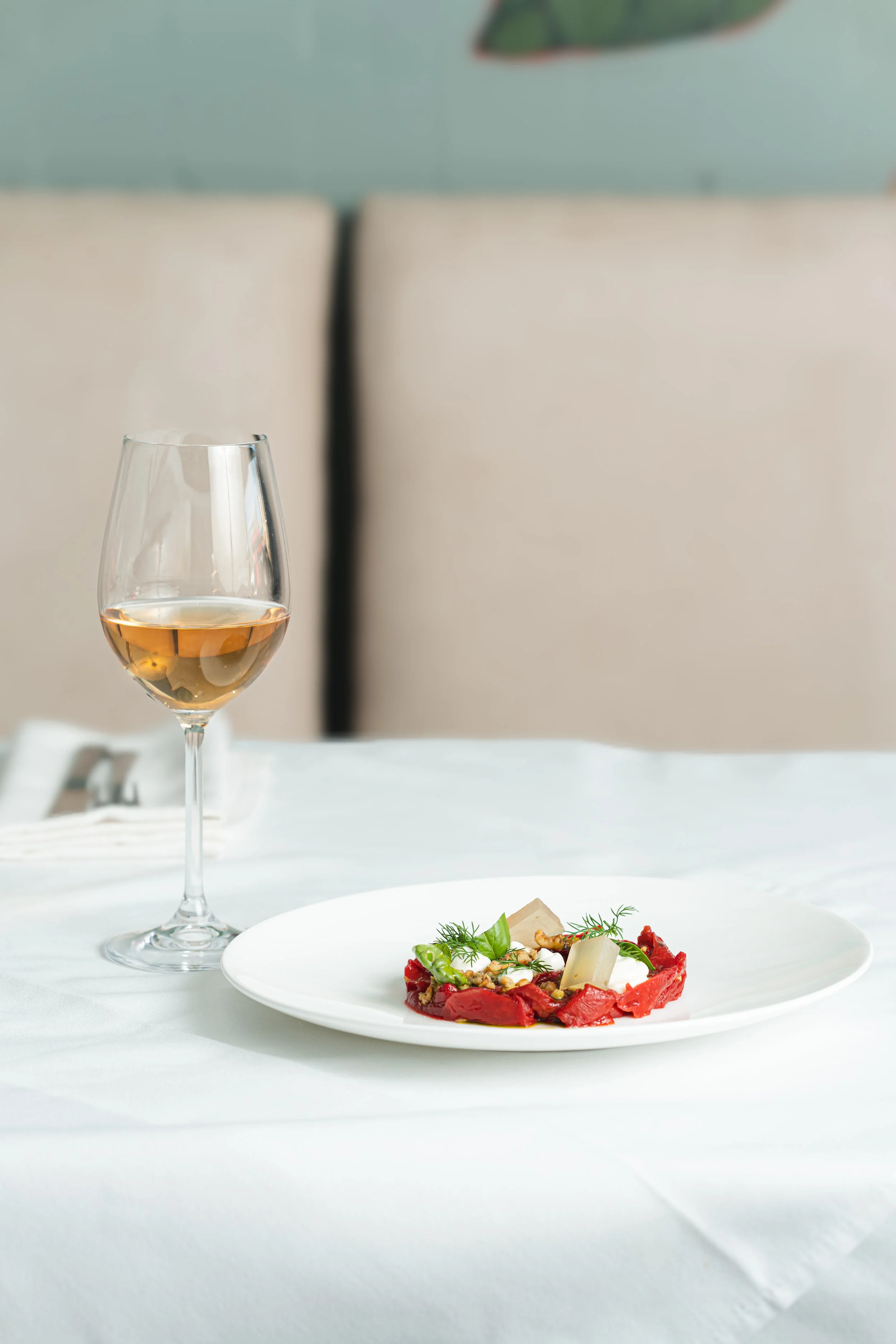 A glass of white wine and a plated appetizer with greens, cheese, and red sauce on a table with a white tablecloth.