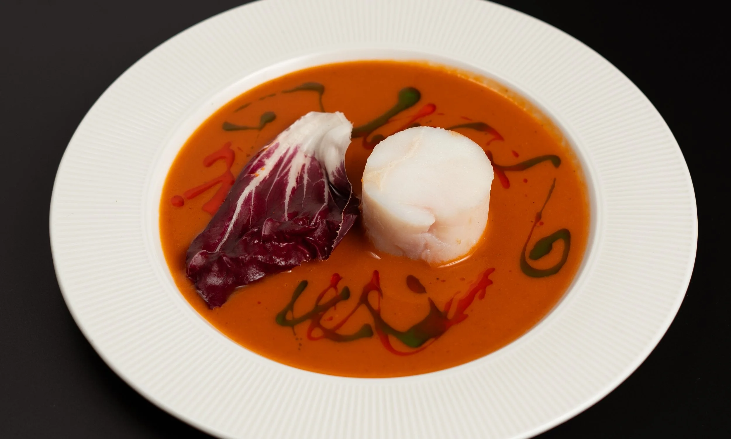 A bowl of tomato-based soup garnished with a leaf of radicchio and a circular slice of white cheese, with green and red sauce or oil drizzled on top.