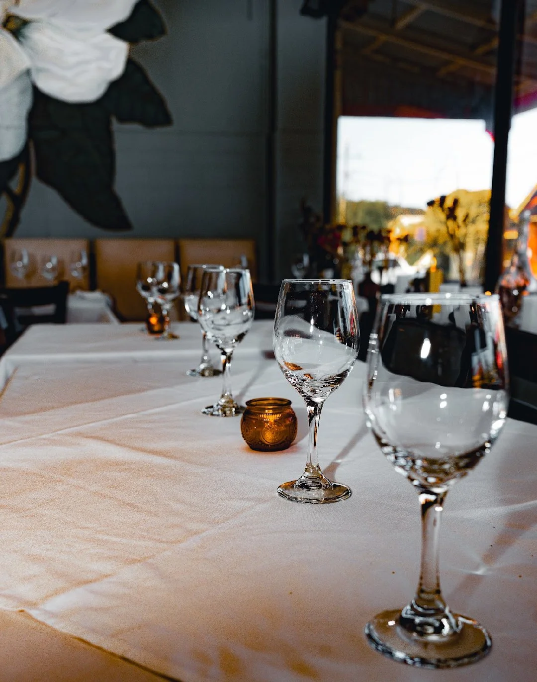 A restaurant table set with empty wine glasses, a small brown candle holder, and a white tablecloth, near large windows with natural light.