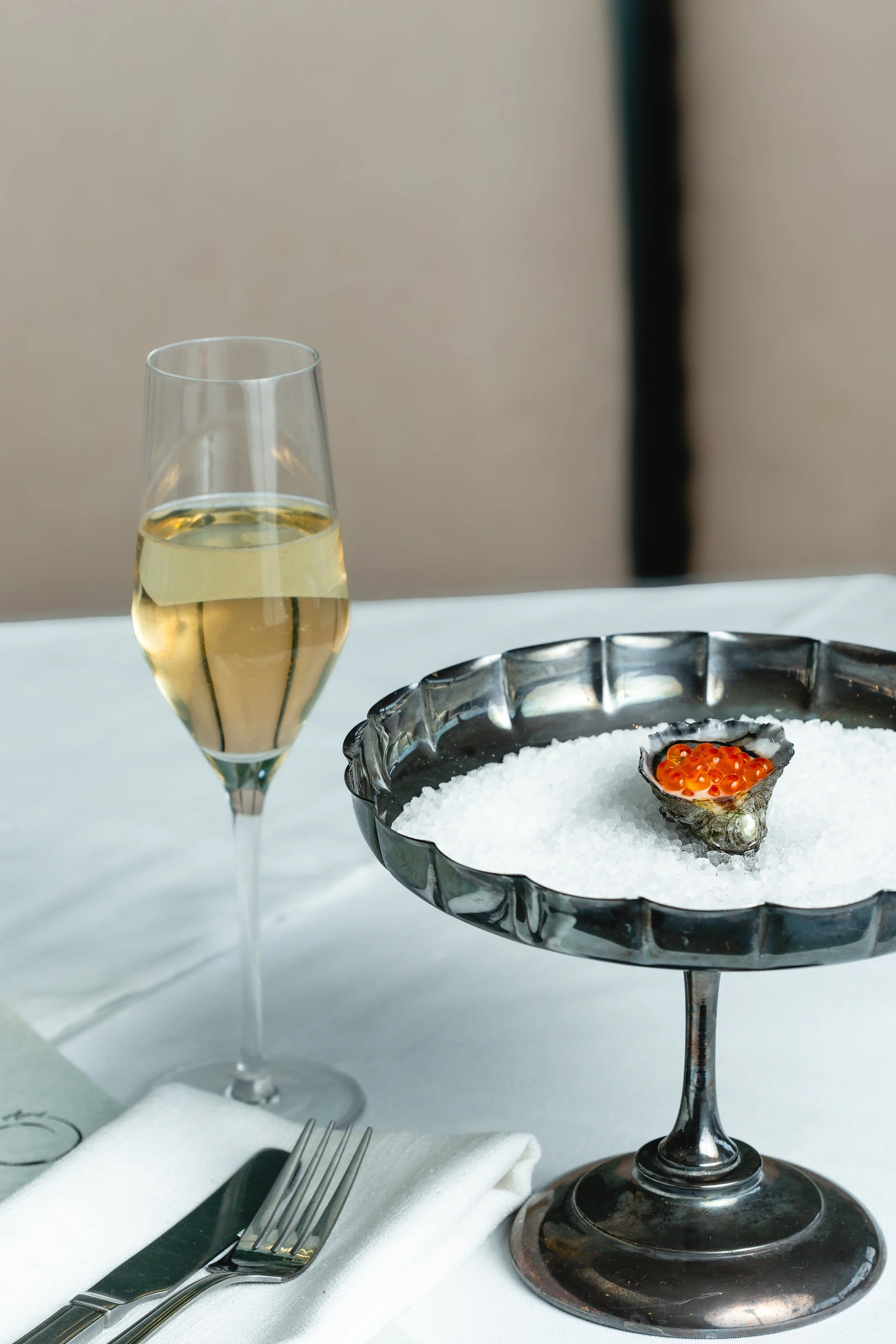 A glass of white wine, a fork and knife on a white napkin, and a silver dish containing salt on a bed of white sea salt with a small black shell filled with red-orange fish eggs, on a white tablecloth.