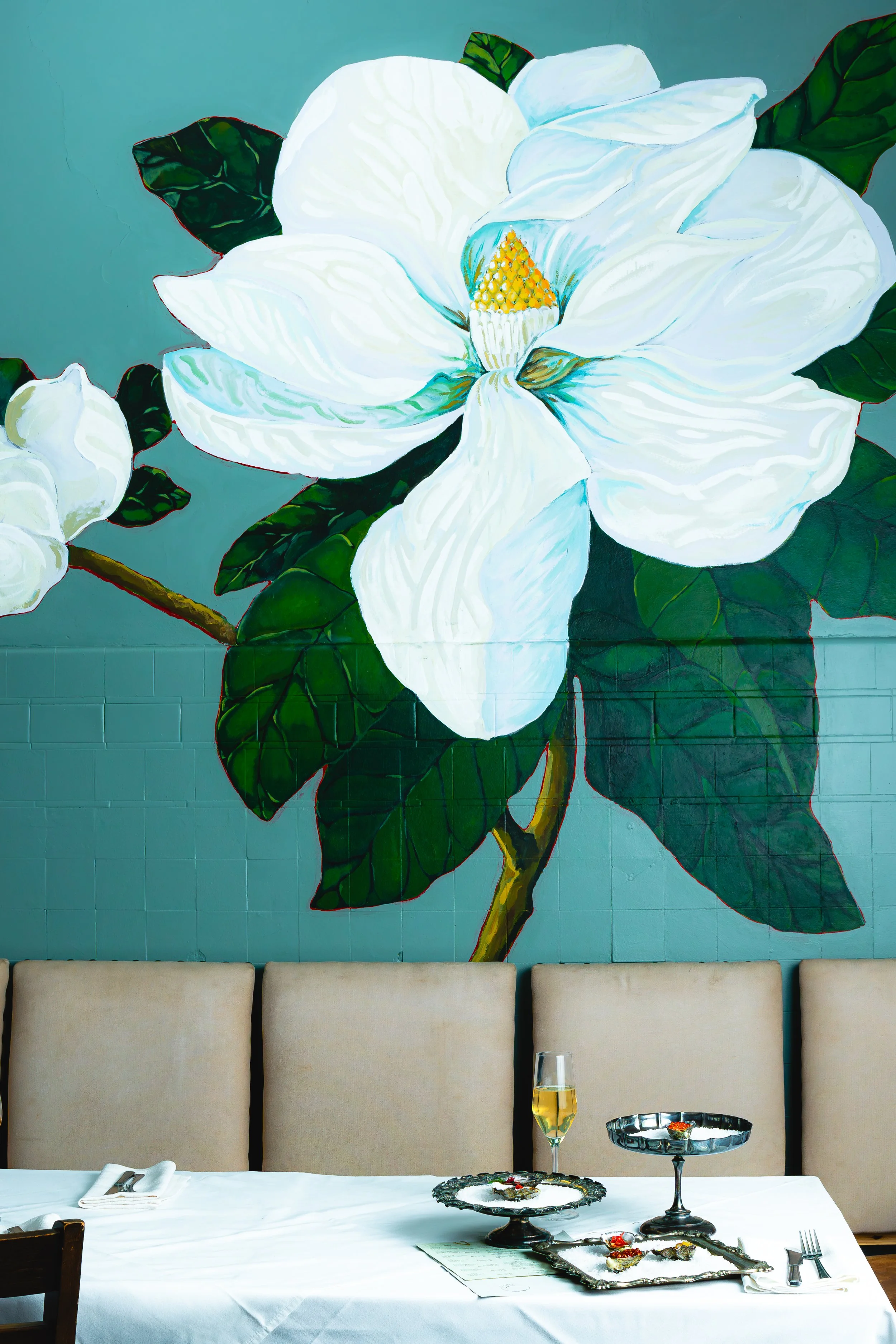 A large white flower painting on a green wall above a dining table with beige chairs, a white tablecloth, a glass of champagne, and decorative serving trays.