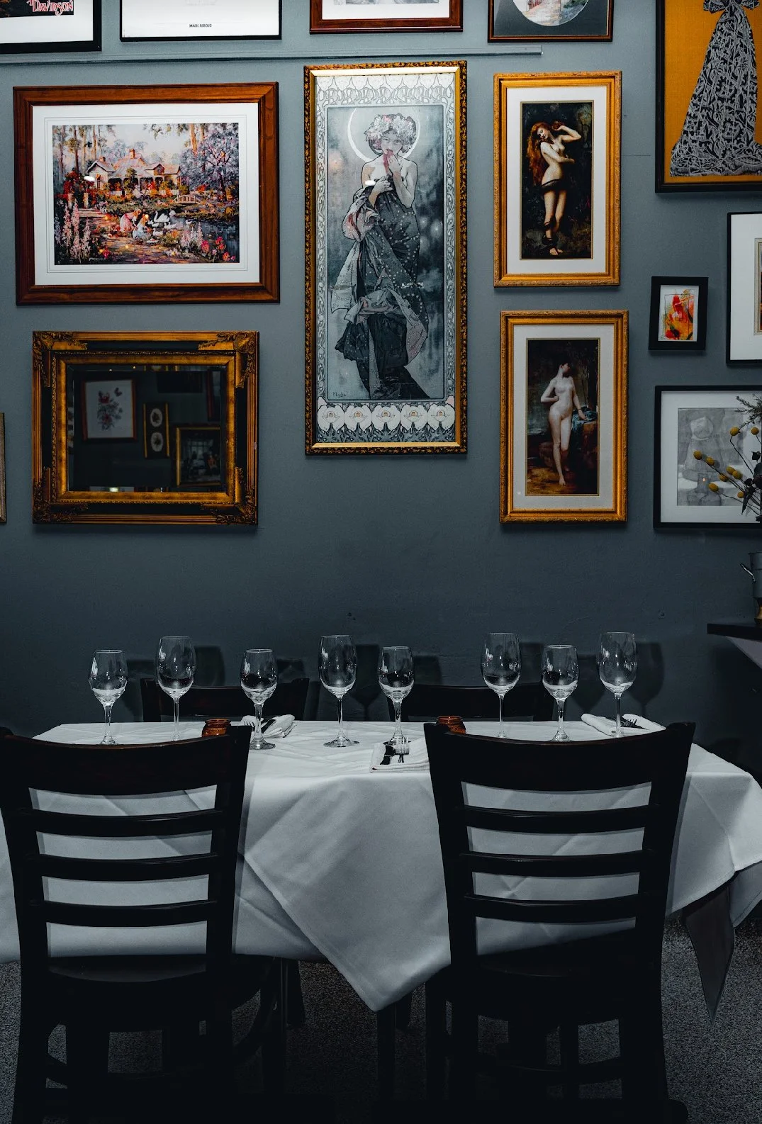 A dining table set for a meal with six wine glasses, silverware, and white napkins in a restaurant with a dark grey wall decorated with framed artwork, including paintings of women and landscapes.