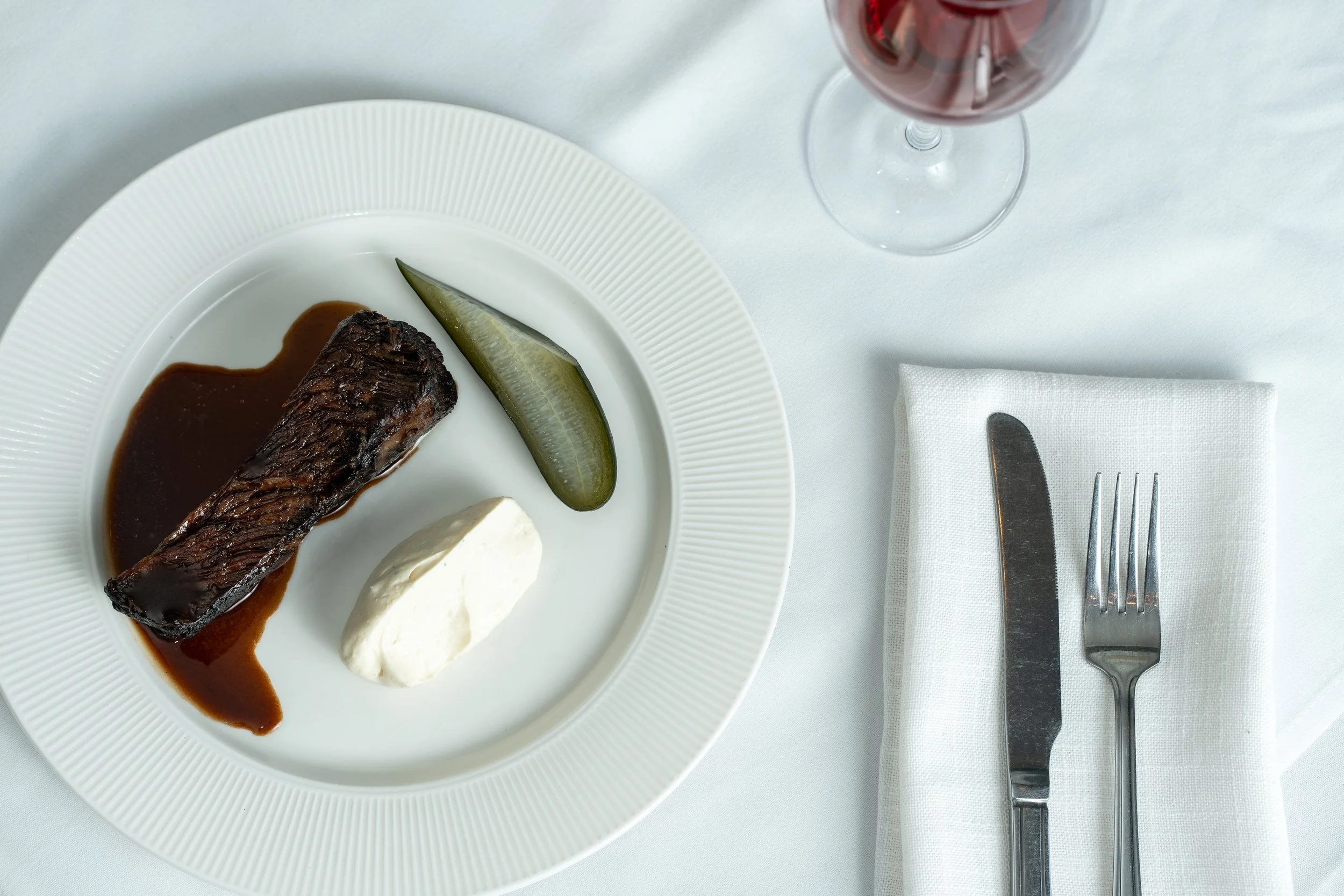 Plate with a piece of braised beef in sauce, a pickle, and a dollop of cream or mashed potatoes, with a glass of red wine, white napkin, knife, and fork on a white tablecloth.