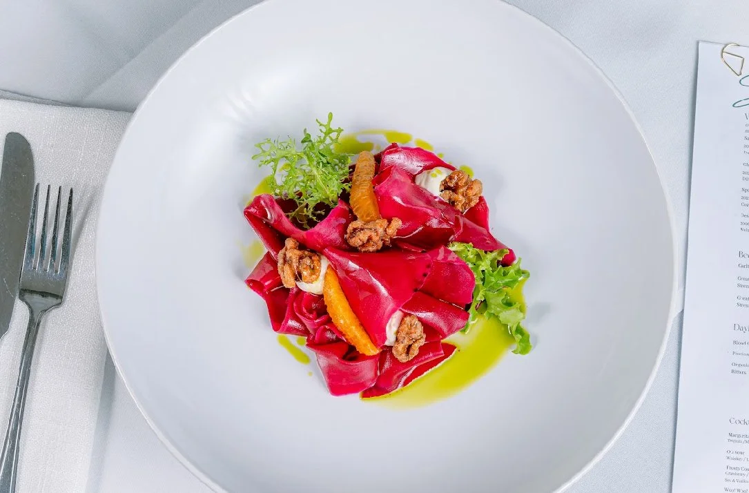 A plated dish with red pasta, garnished with greens, orange slices, walnuts, and a drizzle of yellow sauce, on a white plate at a formal dining setting.