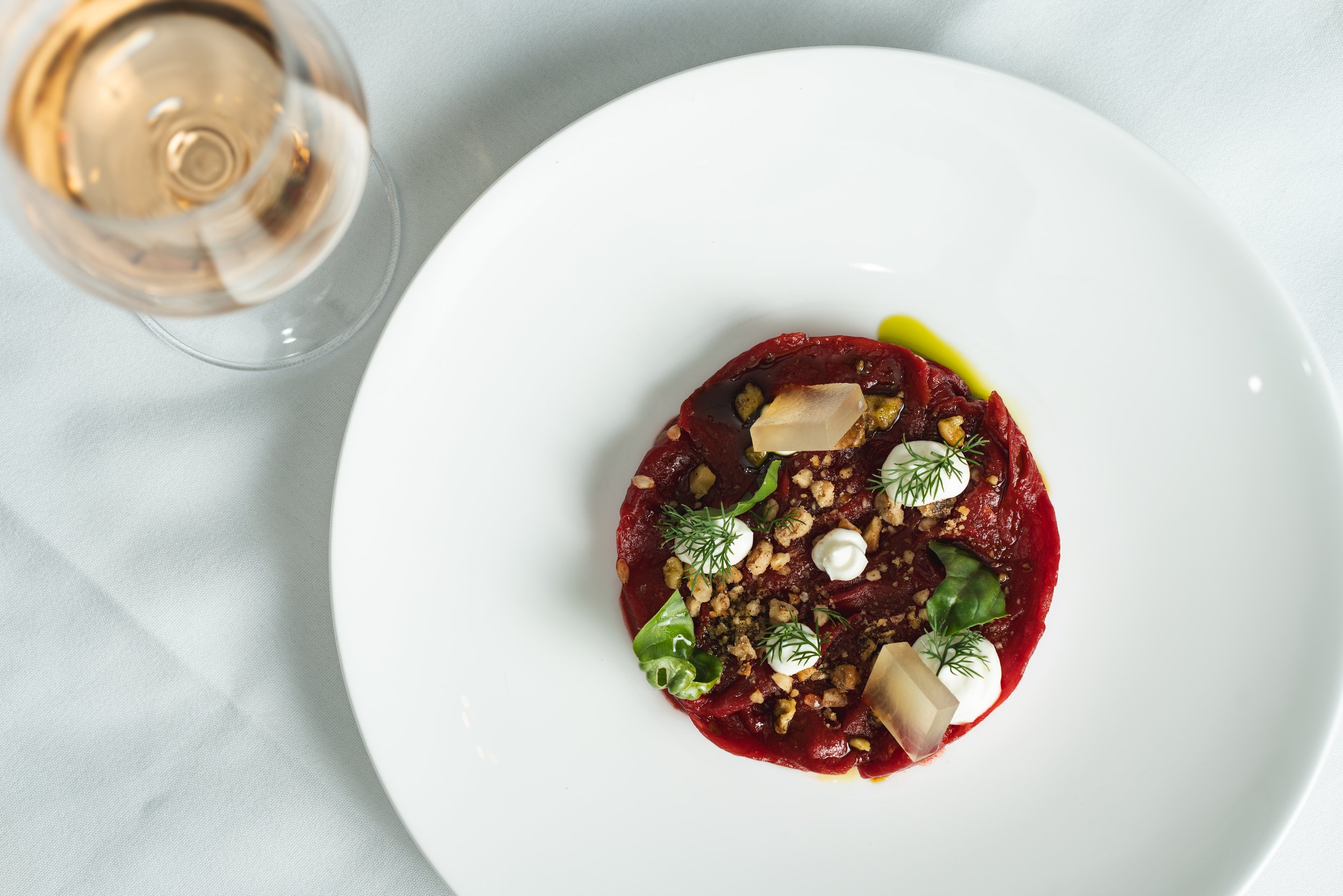 A white plate with a serving of beet carpaccio garnished with microgreens, crumbled nuts, small dollops of white sauce, and small cubes of cheese, with a glass of white wine beside it on a light-colored tablecloth.