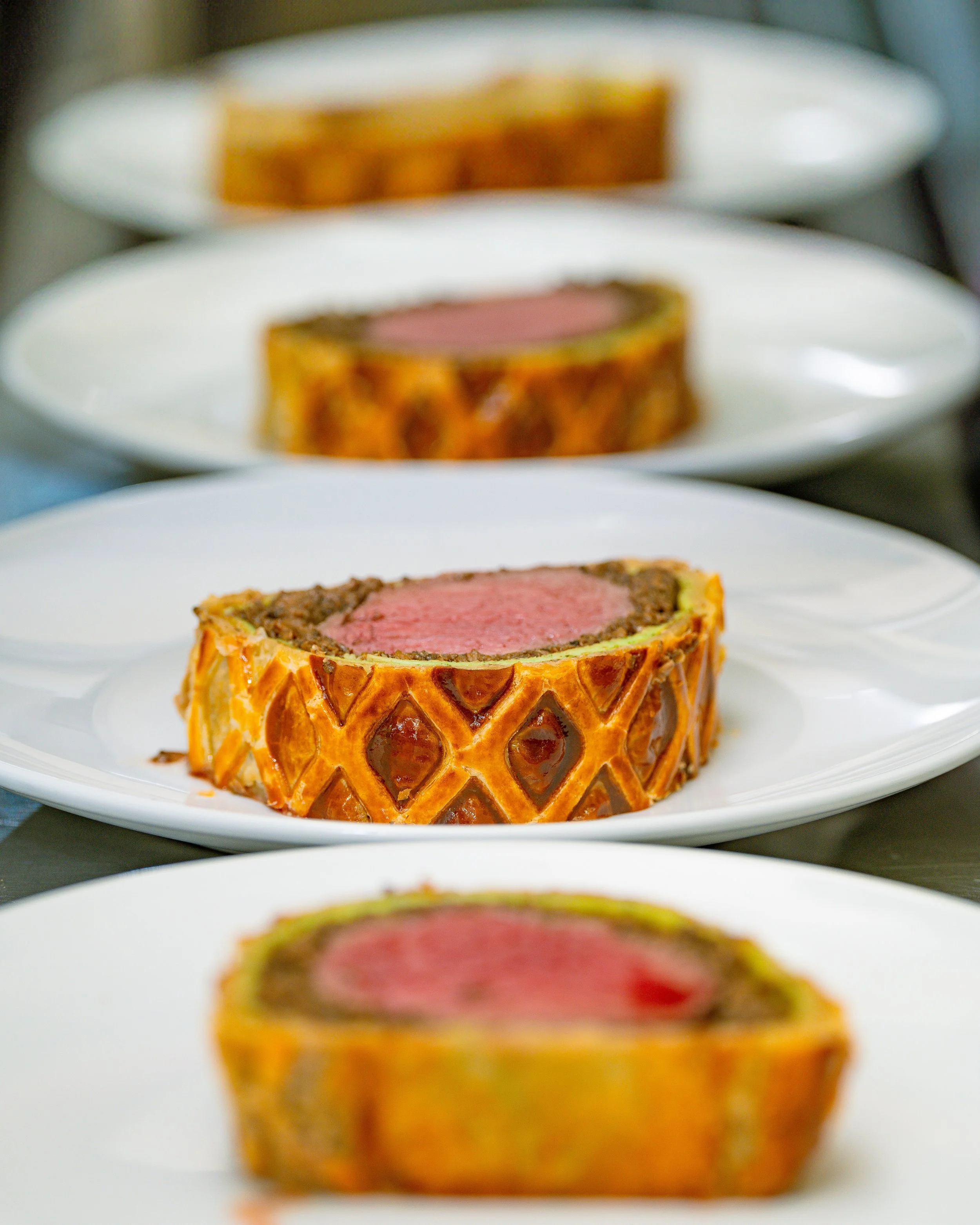 Three white plates with slices of beef Wellington, each showing a pink center, arranged in a line from front to back.