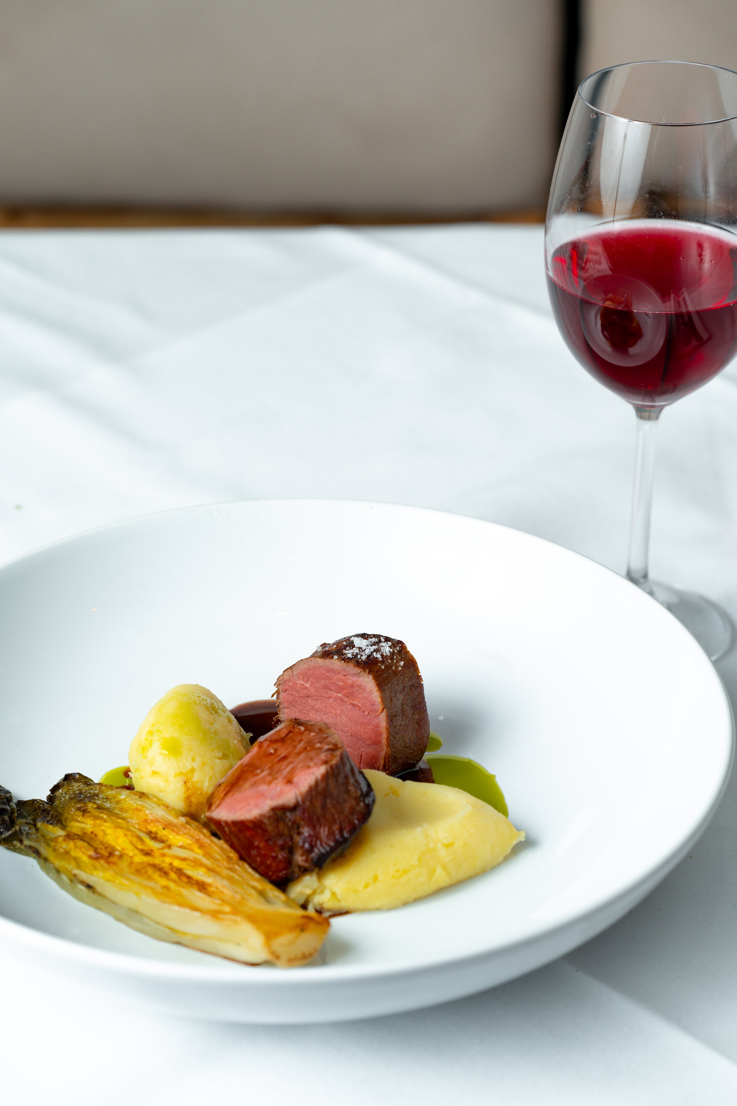Plate with cooked meat, mashed potatoes, vegetable, and sauce next to a glass of red wine on a white tablecloth.