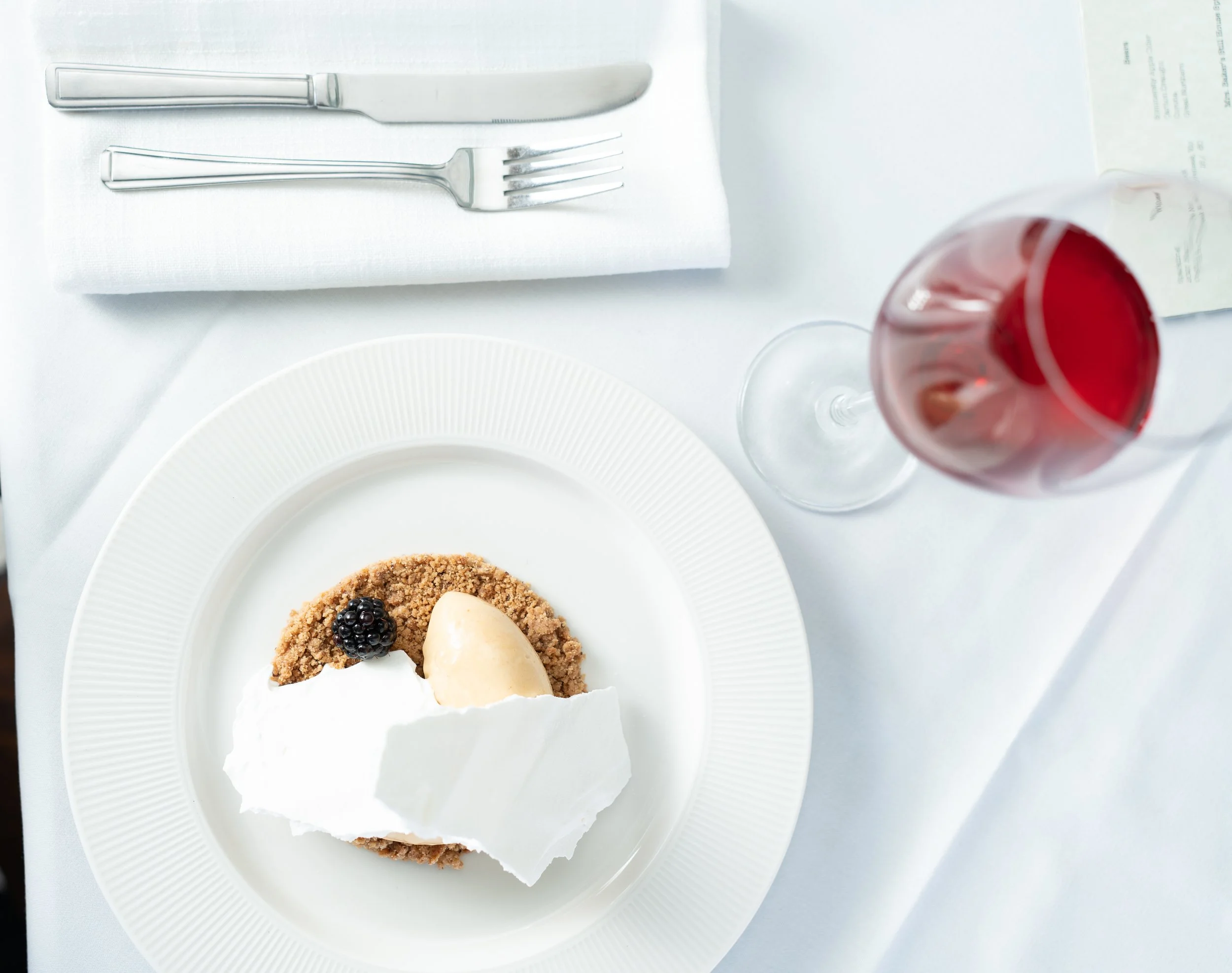 A plate with a slice of dessert topped with whipped cream, a blackberry, and a scoop of ice cream on a white table. There is a glass of red wine beside the plate, with a knife and fork on a napkin in the upper left corner.