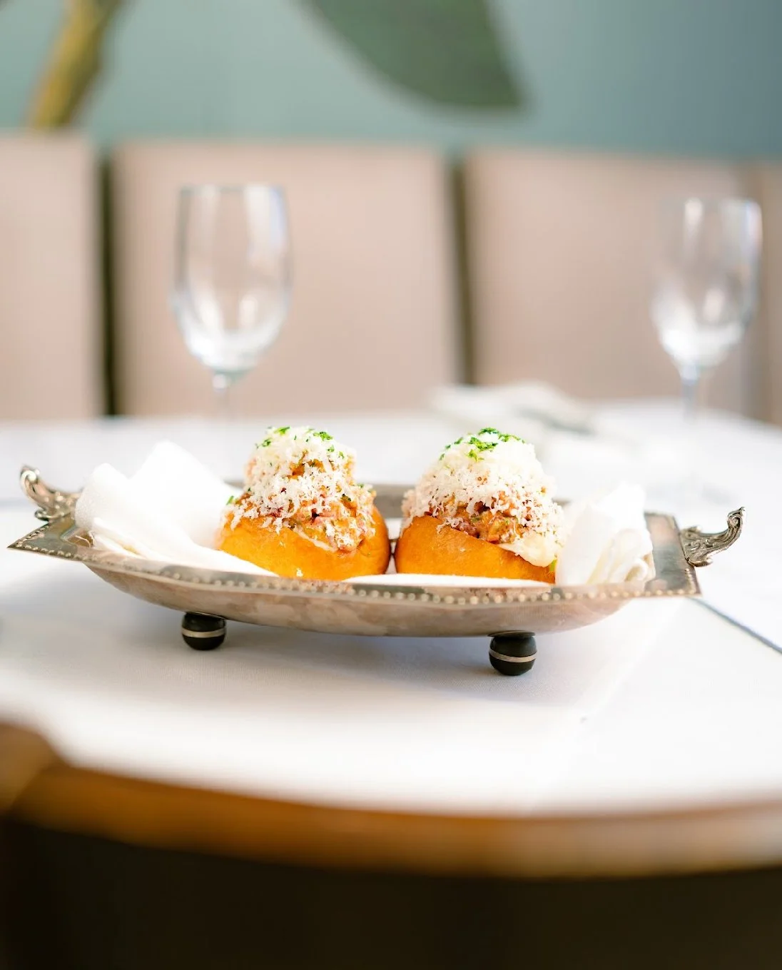 Two stuffed mushrooms with a creamy topping served on a decorative silver platter with dollops of sour cream, on a white tablecloth.