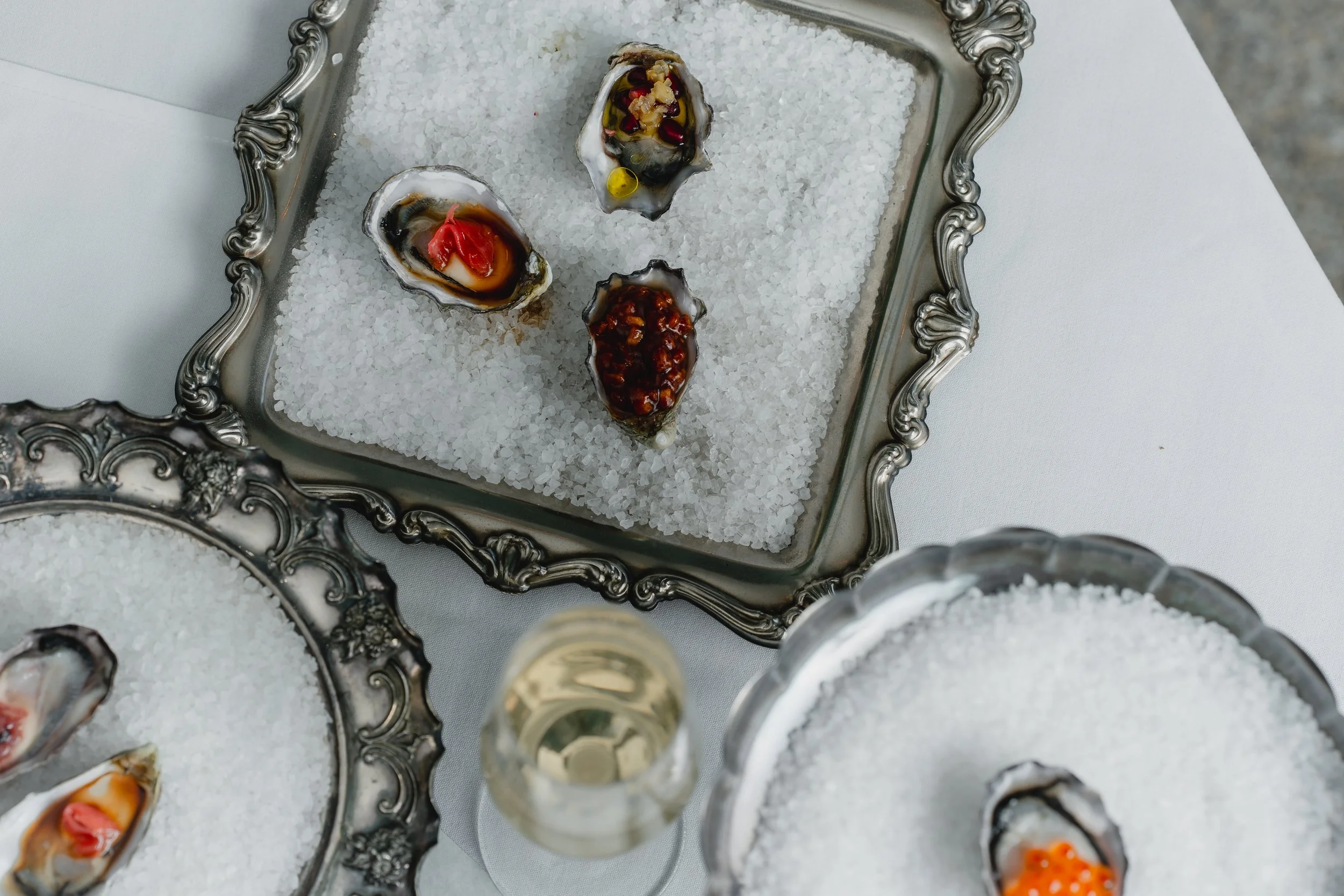 A silver tray filled with ice displaying three oysters topped with various sauces and garnishes. A glass of white wine is partially visible in the foreground, and the tray is placed on a white tablecloth.