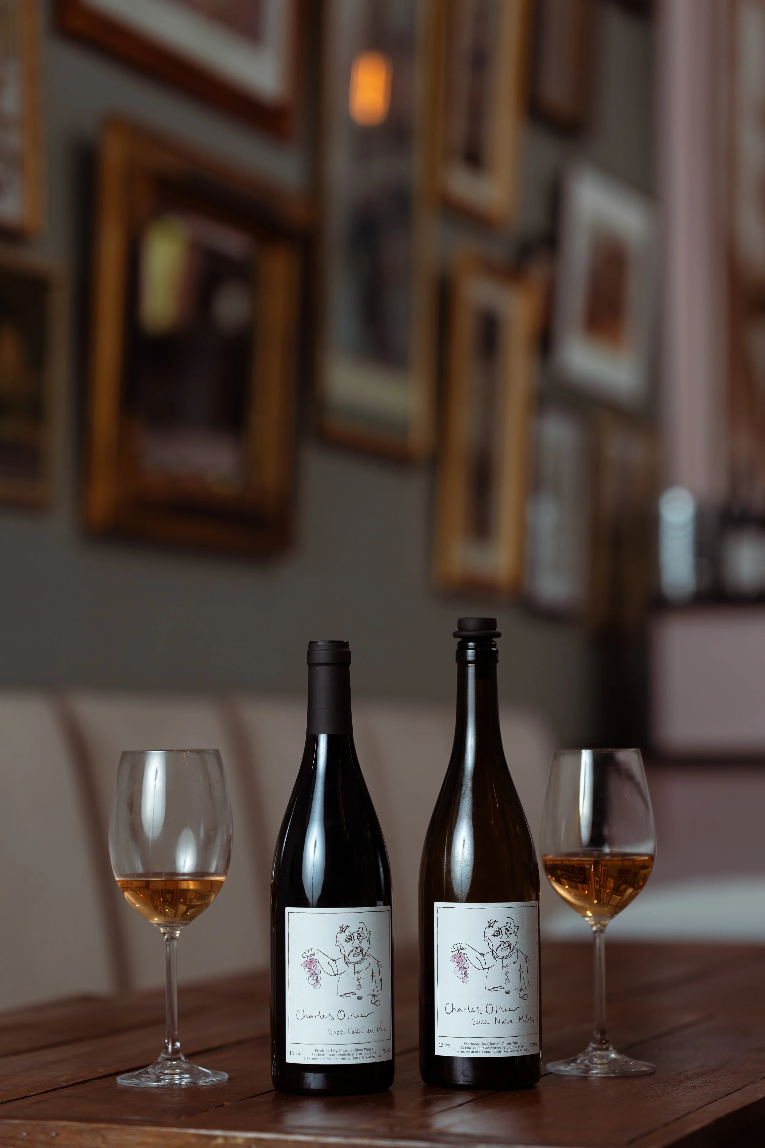 Two bottles of wine with labels featuring a line drawing of a person holding a bunch of grapes, and two glasses of rosé wine on a wooden table in a cozy room with a gallery wall of picture frames in the background.