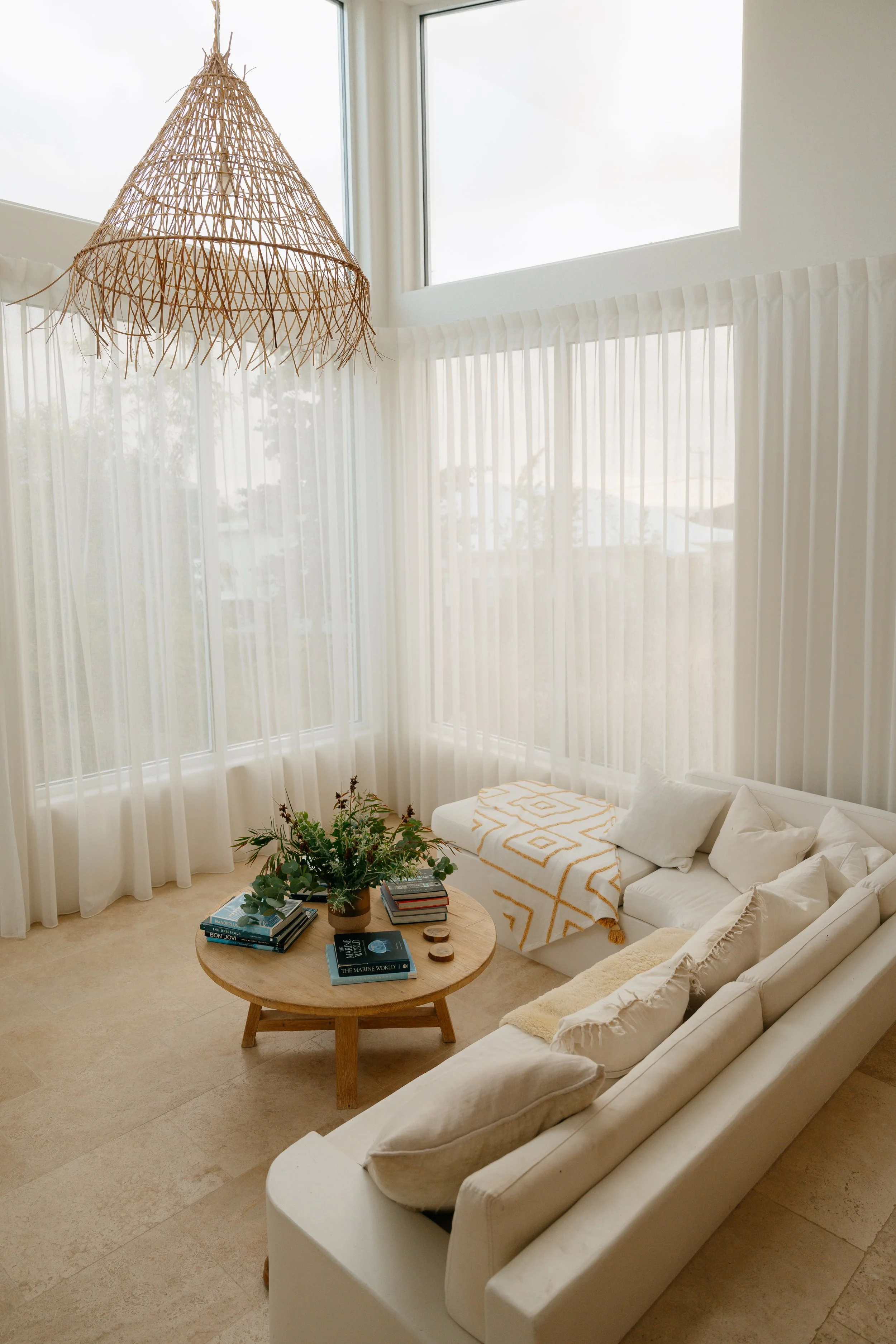 Living room with white curtains, large windows letting in natural light, a white sofa with pillows and a throw blanket, a round wooden coffee table with books, a plant, and candles, and a hanging wicker light fixture.