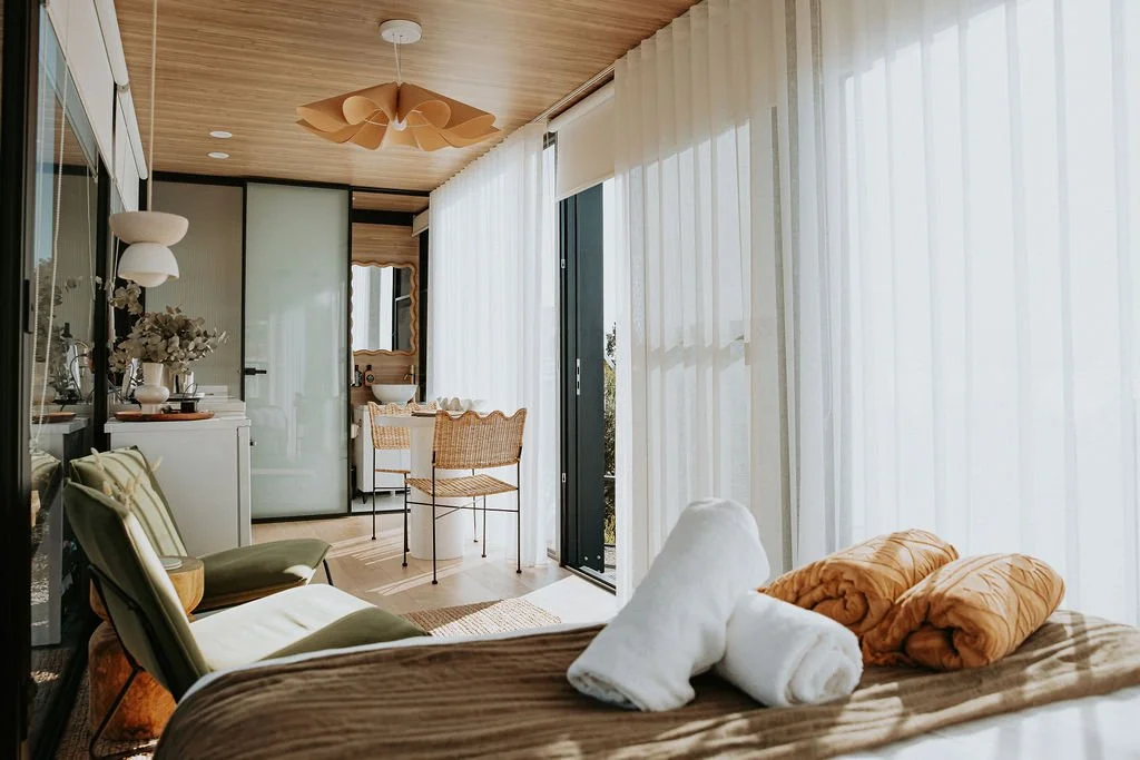 Bright bedroom with large windows and sheer curtains, with neatly made bed featuring towels and blankets, a small sitting area with chairs, a dining table, a bathroom sink, and decorative plants.