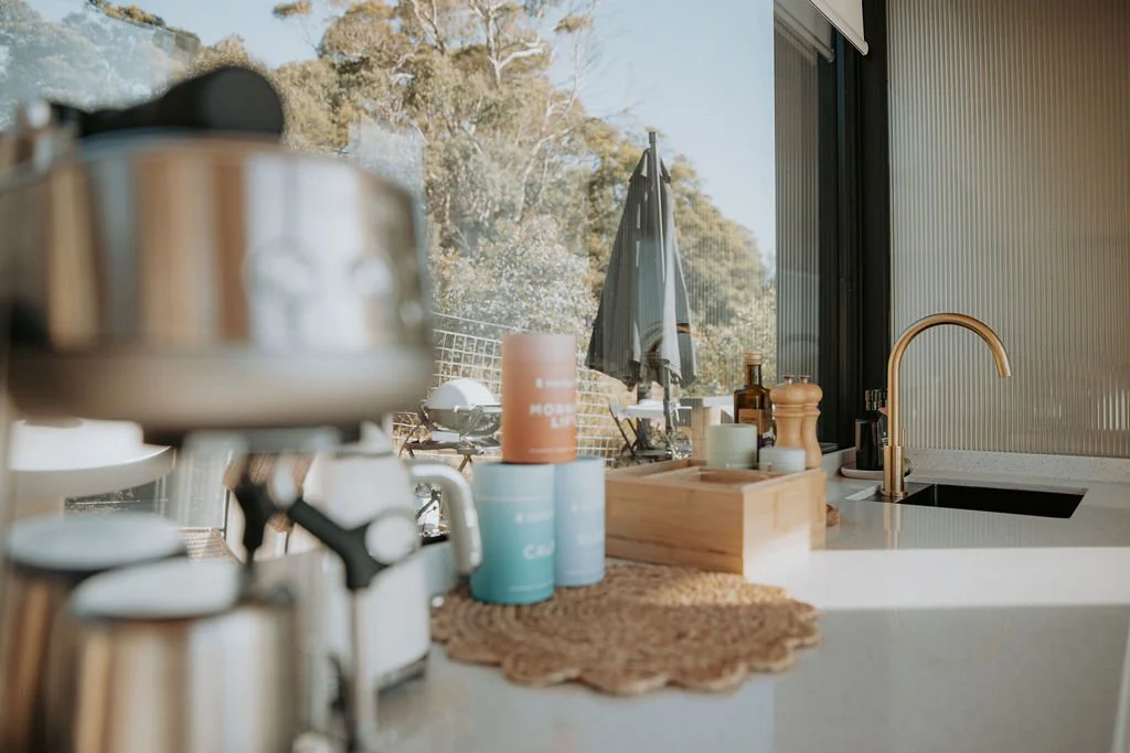 Kitchen counter with coffee cups, a wooden box with soap and lotions, a sink with a gold faucet, and a view of outdoor patio with table and umbrella seen through large windows.