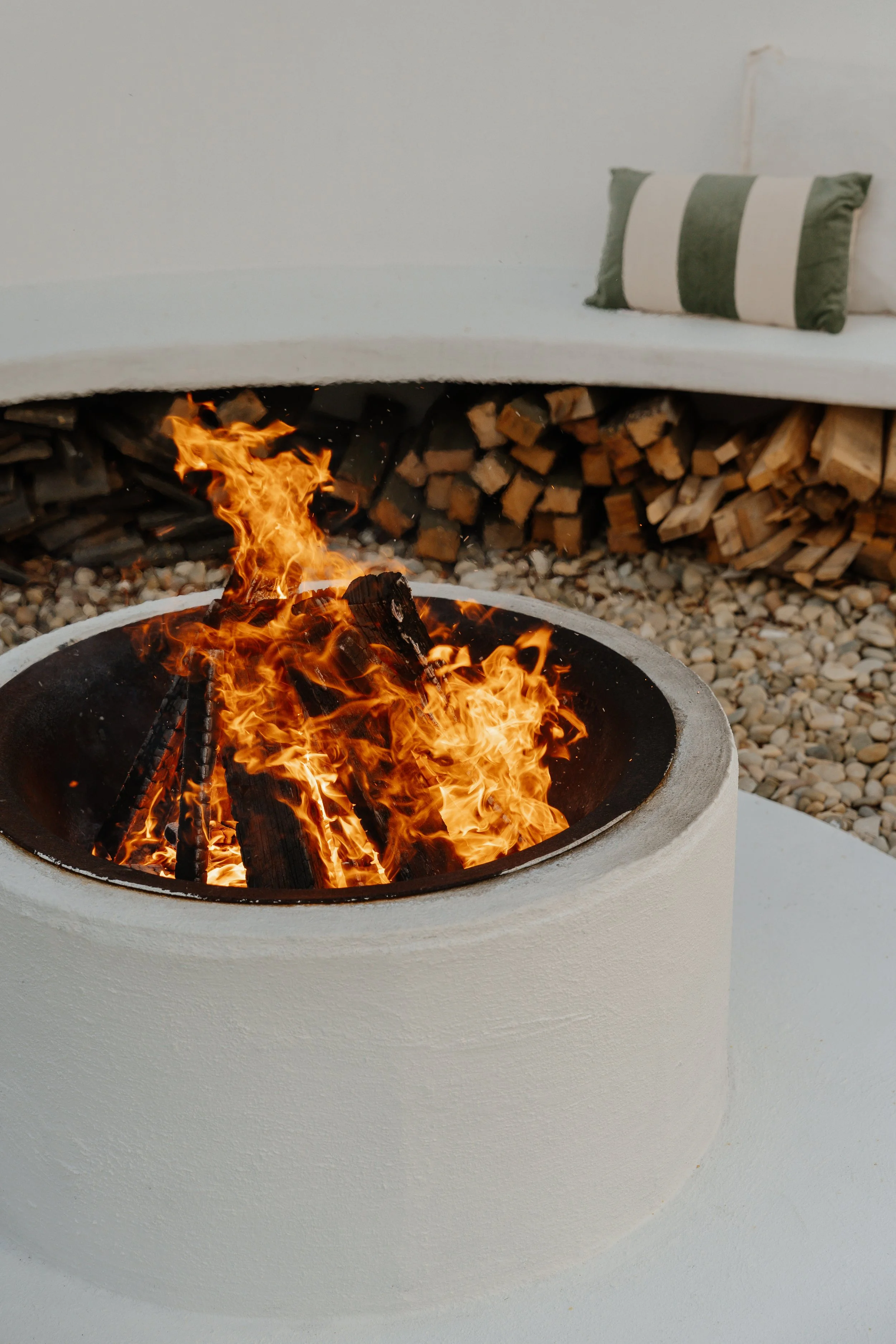 Fire burning in a round outdoor fire pit with wood logs and a white curved seating area with a striped pillow.