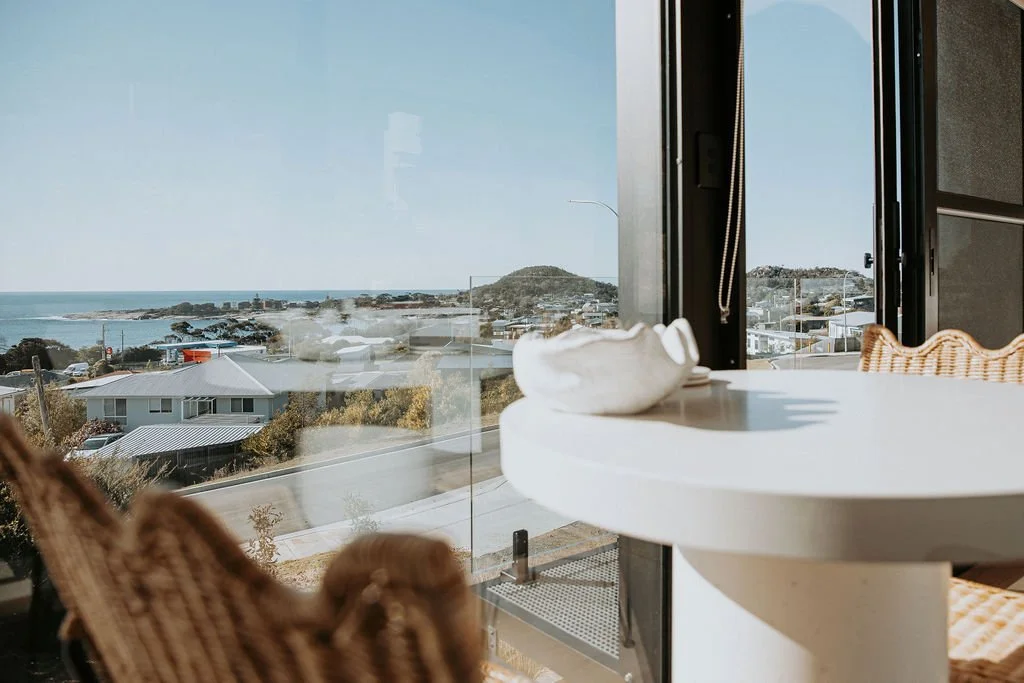 Cozy interior with a round white table, wicker chairs, and large window showing ocean view with houses and distant hills.