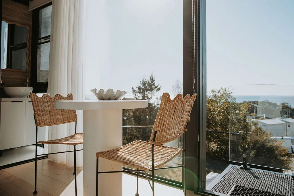 Sunlit indoor space with a round table, two woven chairs, and a view of trees and the ocean through a glass door.