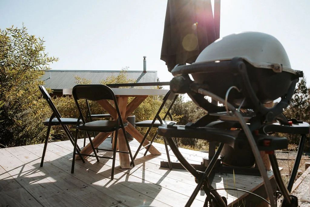 Outdoor wooden deck with a round table and four black chairs, a barbecue grill, and a sunlit scene with trees and a house in the background.