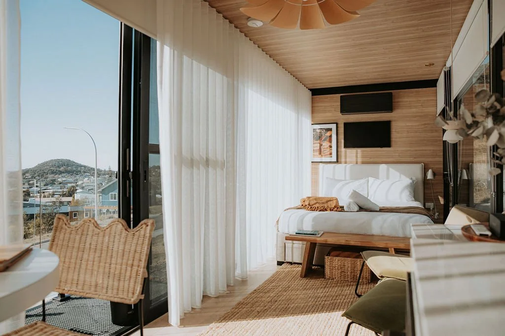 A modern hotel room with a bed, a wooden bench, and sliding glass doors to a balcony with a city view, featuring wood-paneled walls and ceiling, and white curtains.