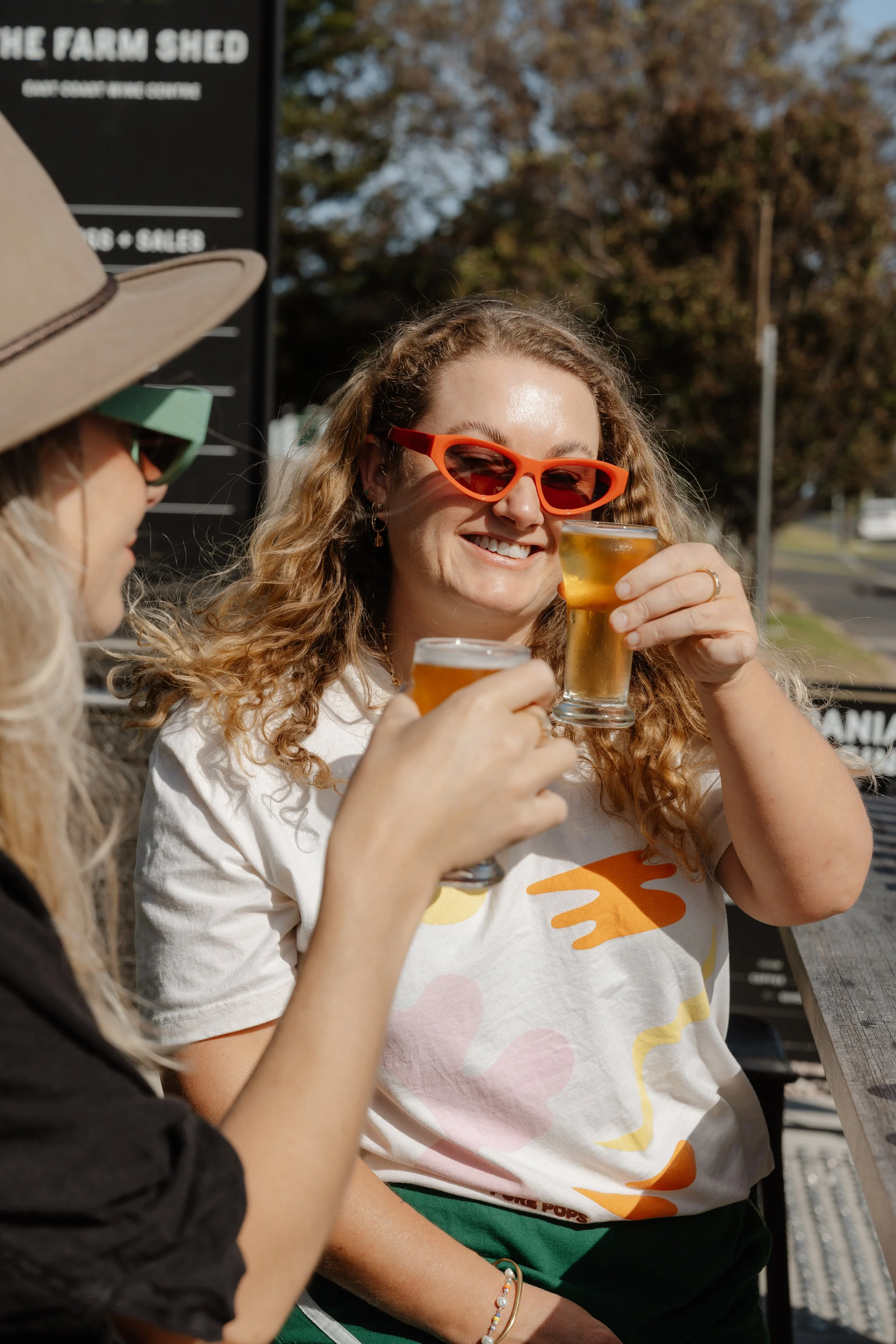 Two women enjoying drinks outdoors. One woman with curly hair and orange sunglasses smiling and holding a glass of beer. The other woman, partially visible, wearing a beige hat and green sunglasses. Background has trees and a sign that reads 'The Farm Shed'.