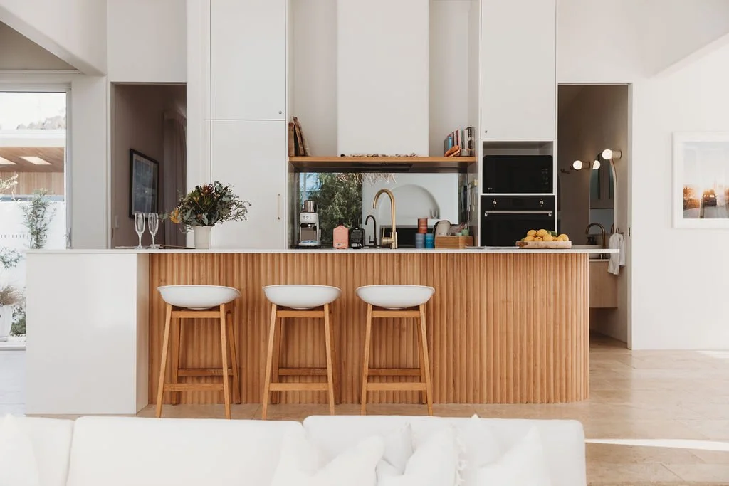 Modern kitchen with white cabinets, wooden accents, and a kitchen island with three white barstools. There is a vase with flowers, a coffee machine, and some fruit on the island.