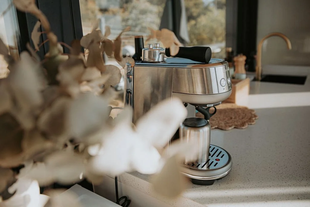 Silver and black espresso machine on kitchen countertop with flowers in foreground and window in background.