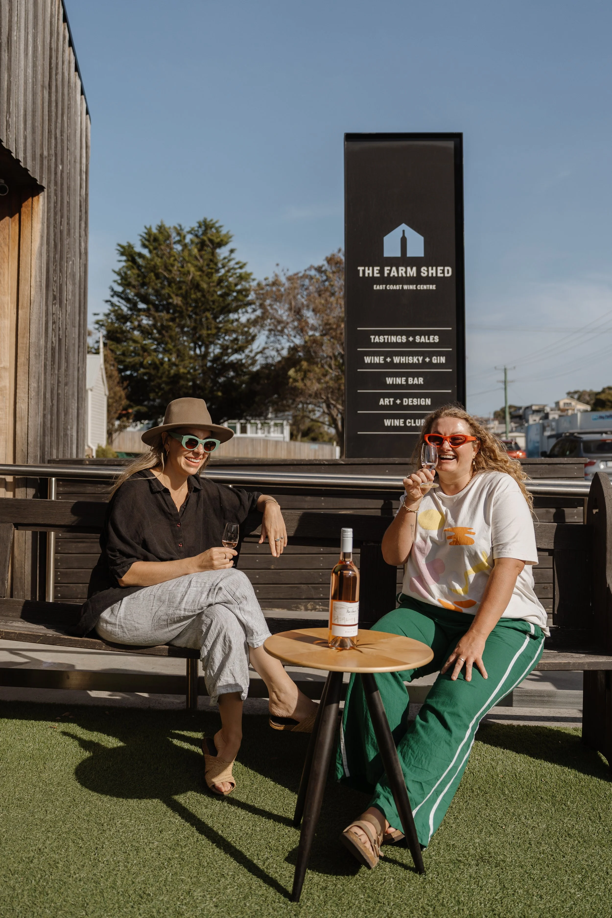 Two women sitting outdoors, smiling and enjoying wine. One woman wears a wide-brimmed hat and sunglasses, and the other has curly hair and orange glasses, holding a wine glass. They are seated at a small round table with a bottle of wine. Behind them is a sign that reads "The Farm Shed, East Coast Wine Centre," listing tastings, sales, wine, whisky, gin, wine bar, art, design, and wine club. There is a rustic wooden building to the left and trees and parked cars in the background.