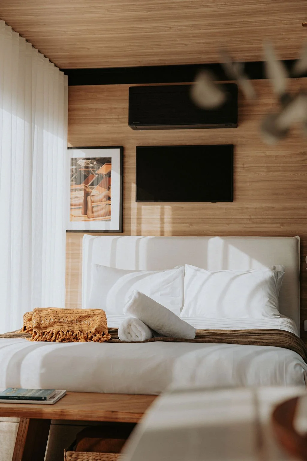 A modern bedroom with a white bed, beige blanket, rolled towels, and framed artwork on a wood-paneled wall with a flat-screen TV and air conditioner above.