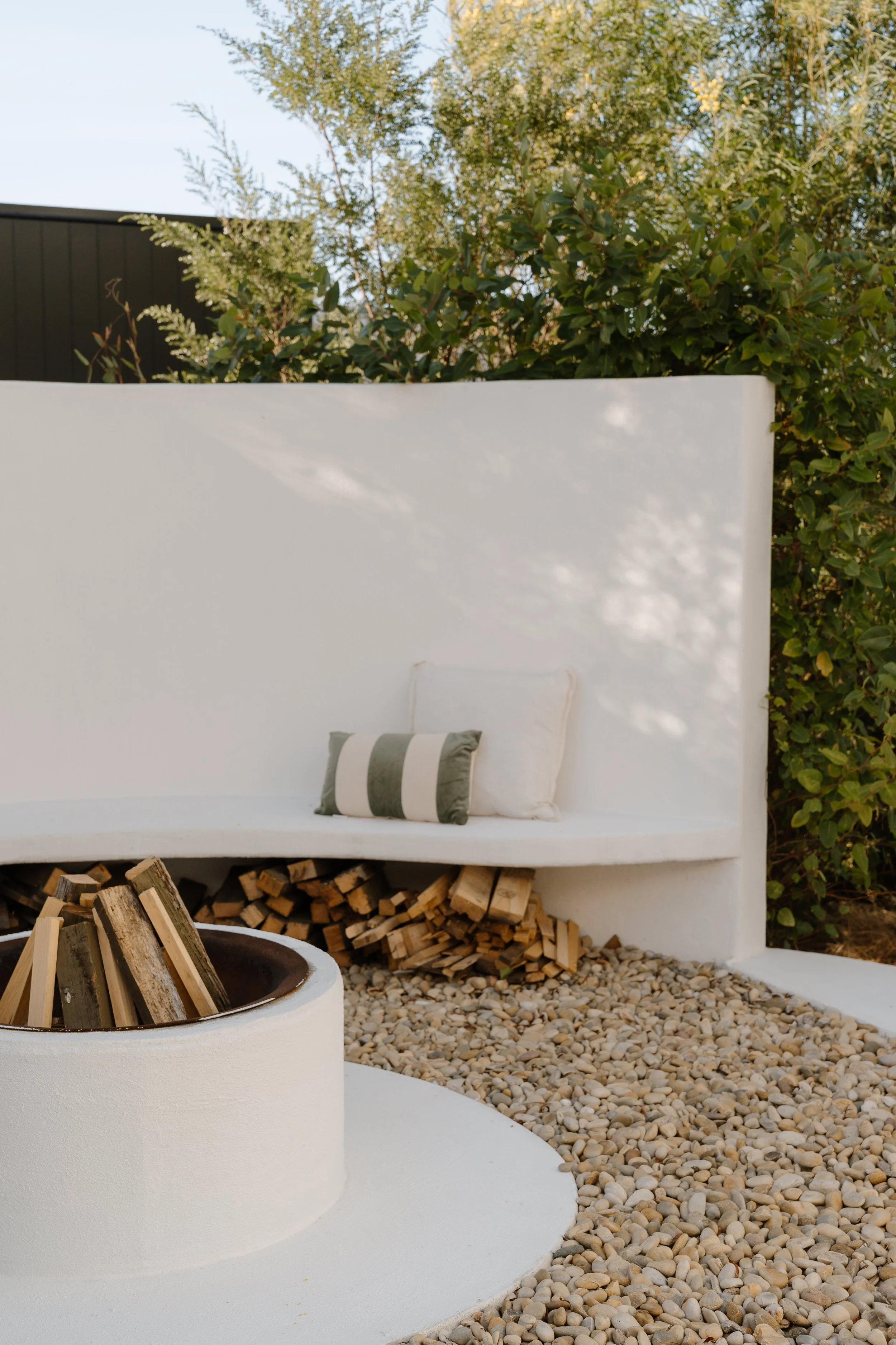 Outdoor seating area with a white wall, a striped pillow, a fire pit with logs, and a gravel ground, surrounded by greenery.