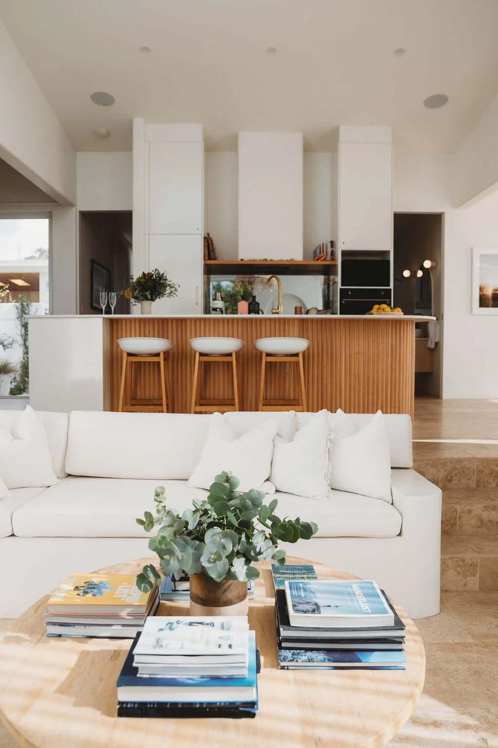 Modern open-concept living room and kitchen with white sofa, wooden coffee table with magazines and a plant, and a kitchen island with three white bar stools, a gold faucet, and pendant lighting