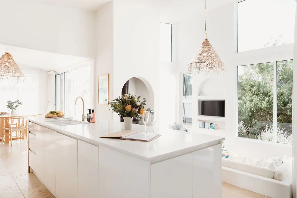 Bright, modern open-concept living space with white walls, large windows, and minimal decor, including a kitchen island with flowers and wine glasses, and a sitting area with a white sofa.