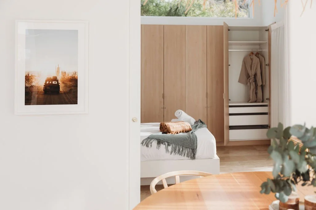 Part of a bedroom with a bed covered with a blanket and pillows, a wardrobe with an open door showing hanging clothes, a wall with a framed sunset or desert landscape photograph, and a dining table with a plant in the foreground.