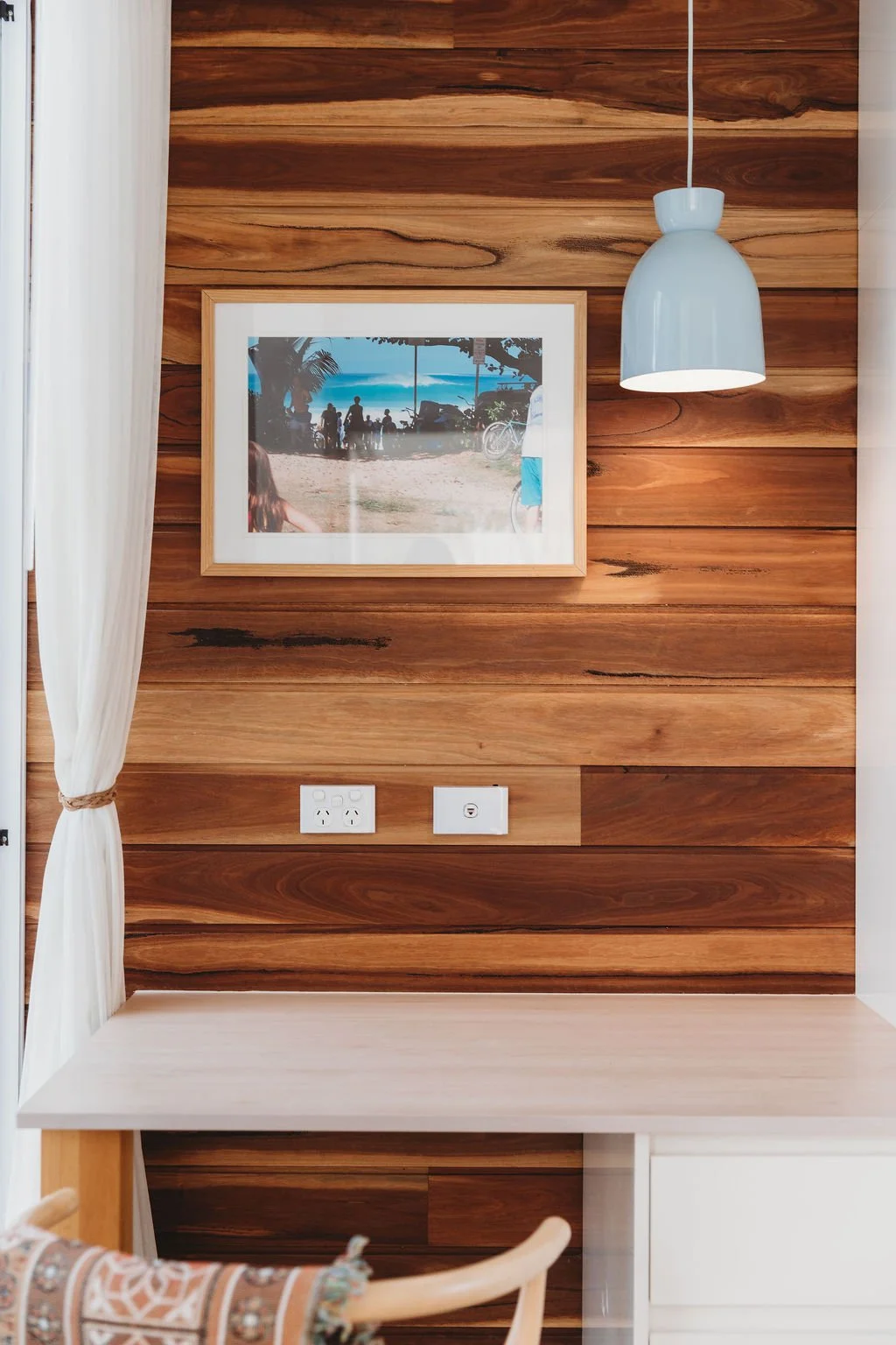 A wooden wall with a framed beach photograph, a modern white hanging lamp, two electrical outlets, a white desk, and a light-colored curtain on the left side.