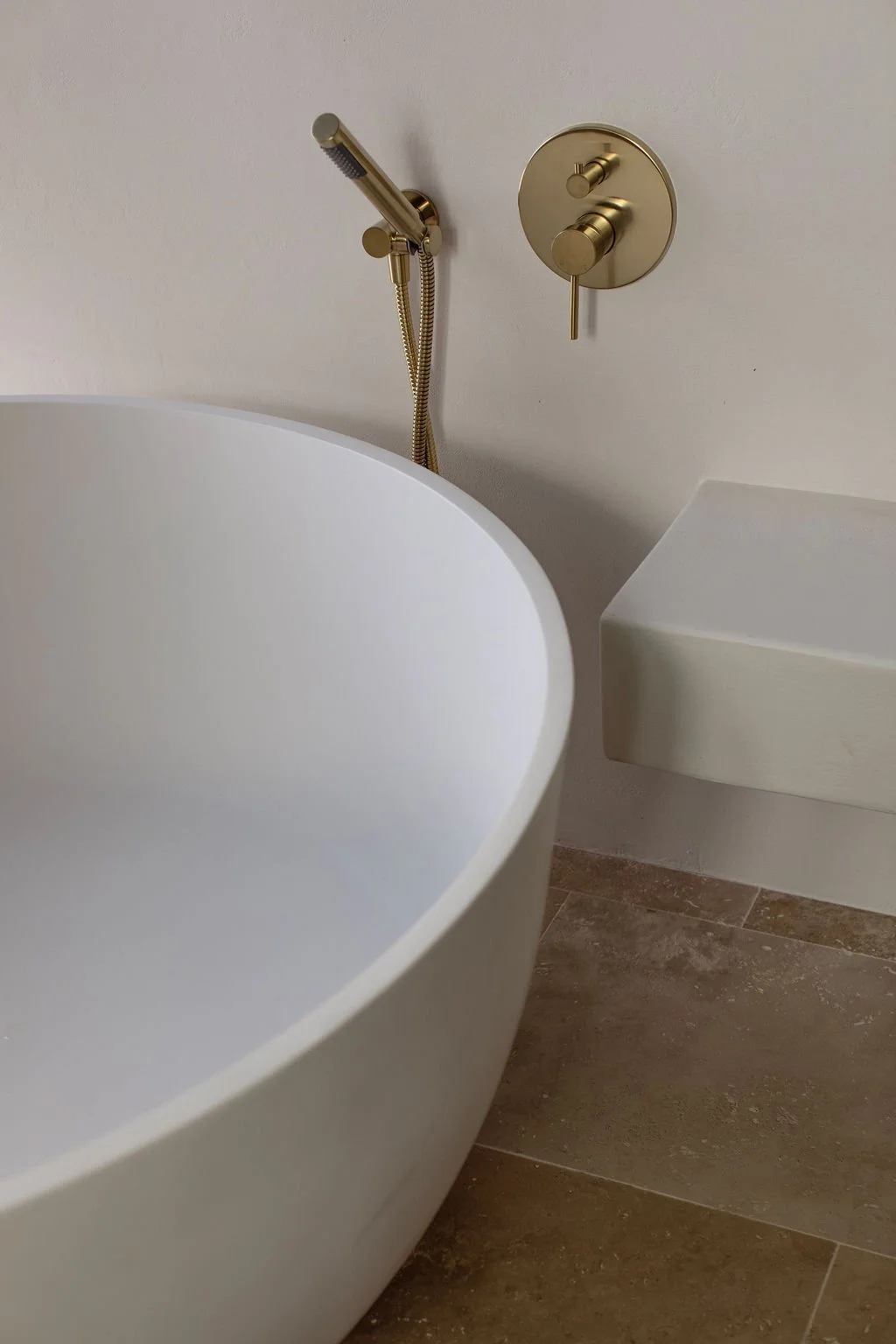 Close-up of a modern bathtub, with a gold-colored handheld shower and a matching shower control on a white wall, next to a beige tiled floor and a white ledge.