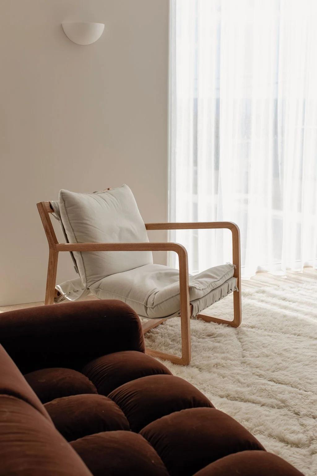 A modern living room corner with a beige cushioned wooden armchair, a dark brown velvet sofa, a white shaggy rug, and sheer white curtains letting in natural light.