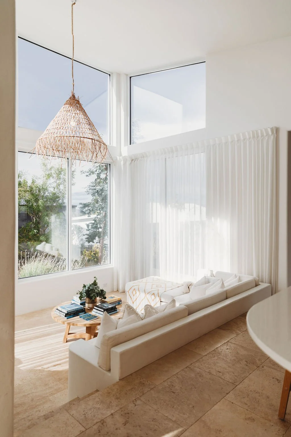 Bright living room with large windows, white curtains, a white sectional sofa, a round wooden coffee table with books and a plant, and a woven pendant light.