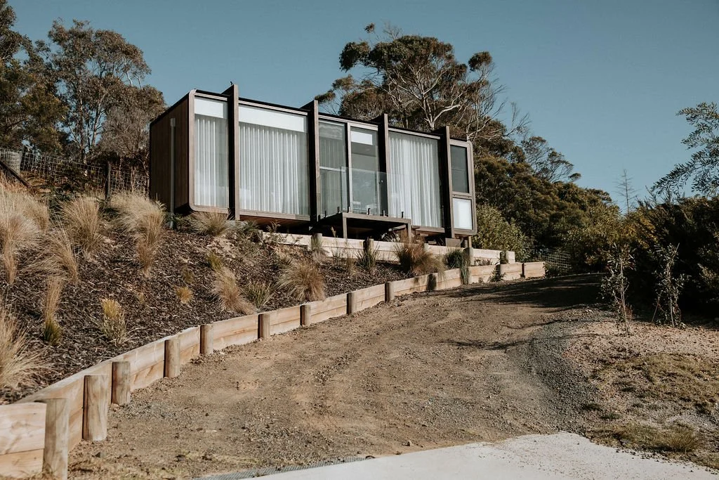 Modern house with large glass windows and a balcony, built on a hillside with terraced landscaping, surrounded by trees and shrubs.