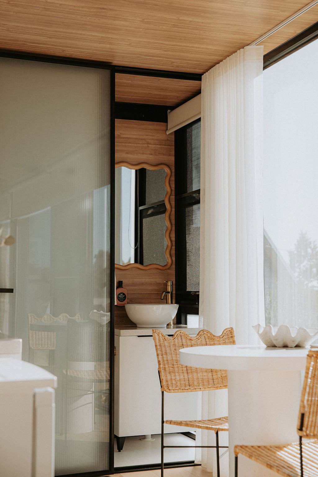 Interior of a modern room with a small bathroom vanity, mirror, and wicker chairs around a white round table, with large window and white curtains.